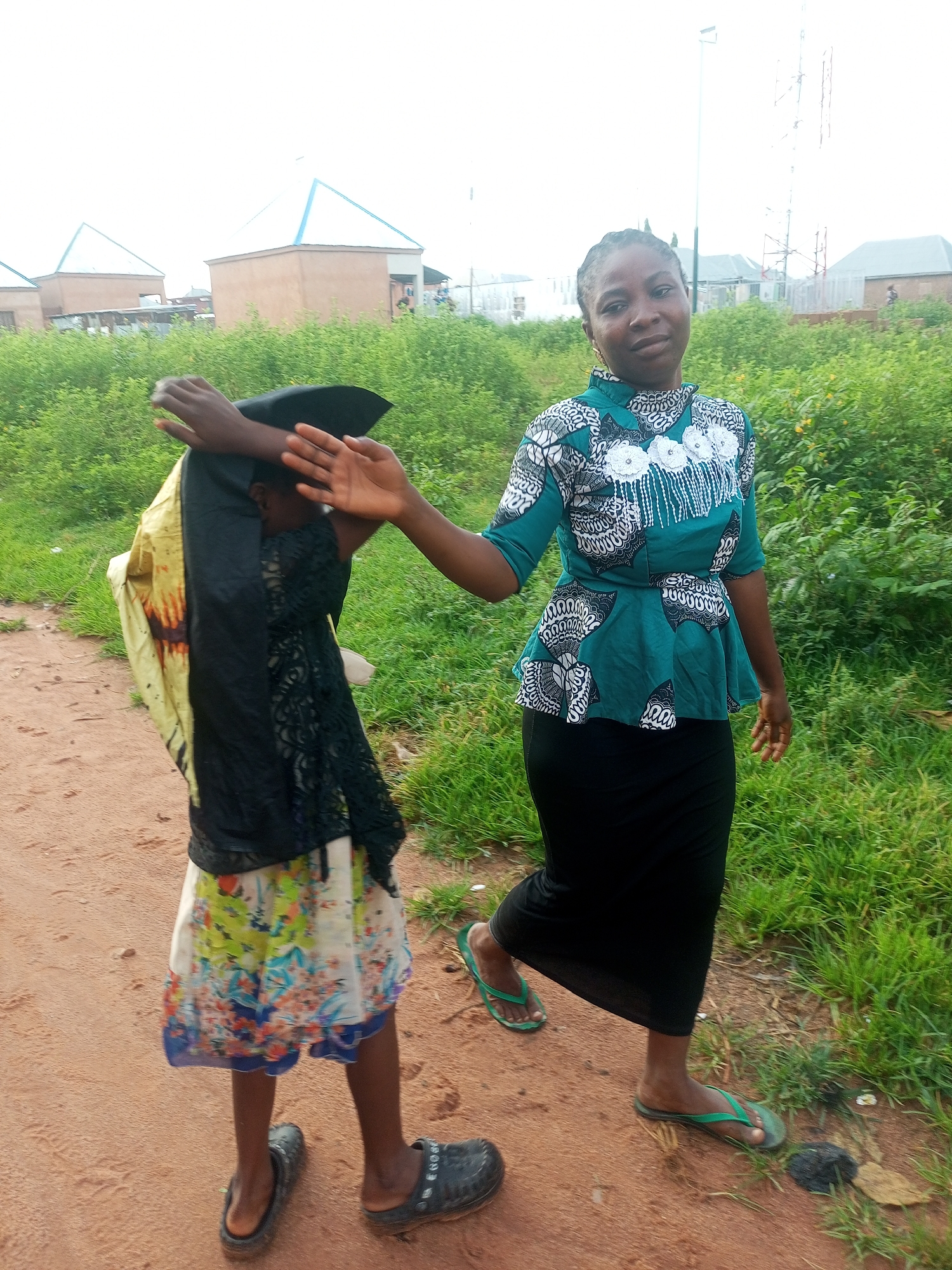 The image depicts two individuals on an unpaved dirt road in Tempure, Nigeria. The person on the right is an adult female wearing a teal and white patterned peplum top and a black skirt, with green flip-flops. She is facing the camera and looking towards it. To her left, a child is partially obscured by a black and yellow garment draped over their head and shoulders. The child is wearing a colorful, patterned skirt and dark clogs. Their hands are raised, with one covering their face and the other extended outward. The environment includes lush green vegetation lining the right side of the road, and in the background, several buildings with peaked roofs are visible. A tall communication tower stands in the distance to the right. The overall lighting suggests it is daytime, with an overcast sky.