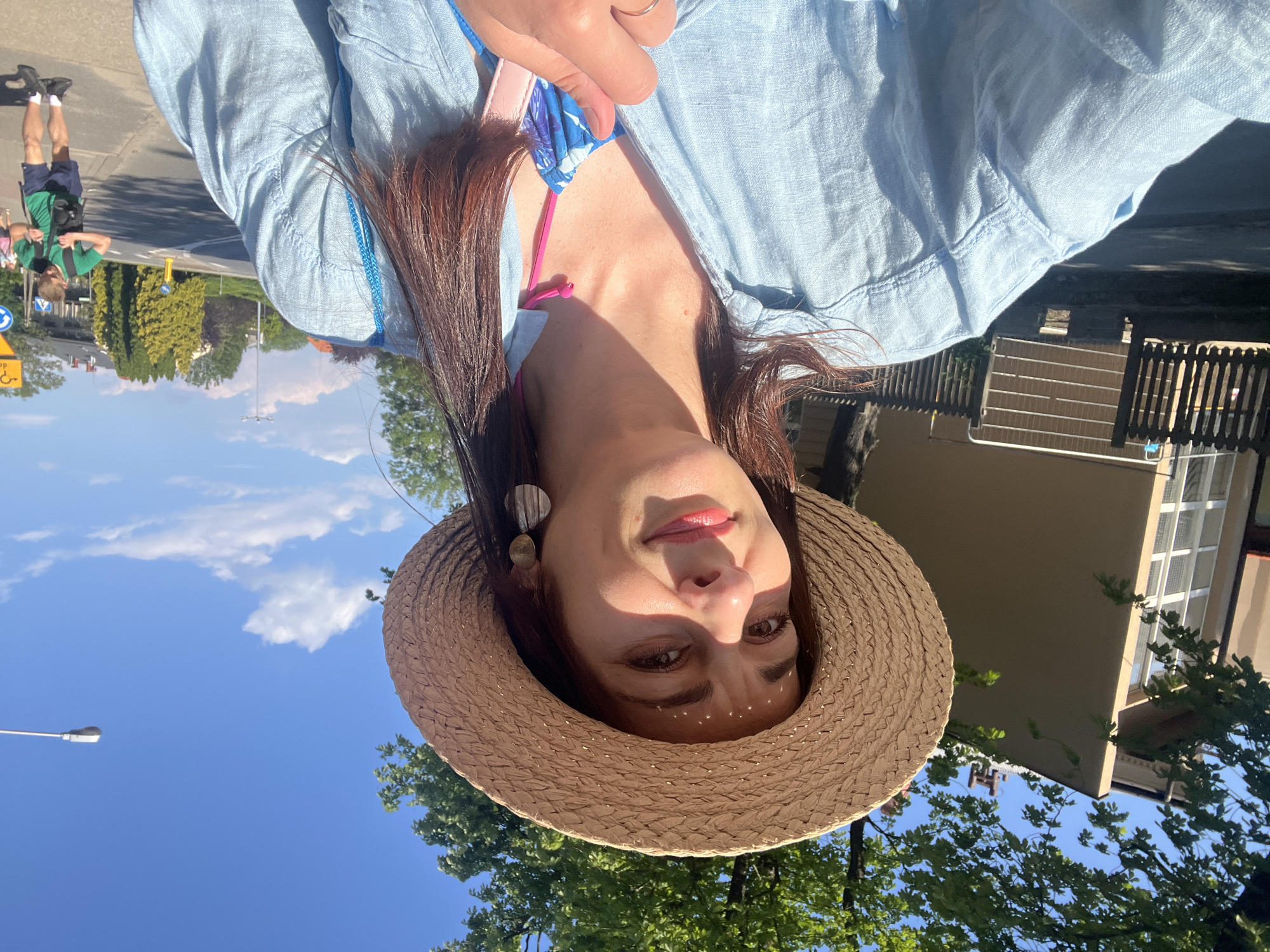 A woman wearing a straw hat and a light blue shirt takes a selfie outdoors on a sunny day. She has long, dark hair and is wearing earrings and a ring. A bright blue bag with a pink strap is visible. In the background, there's a building with a fence, trees, a street with a yellow road sign, and a few people walking. The sky is blue with some white clouds.