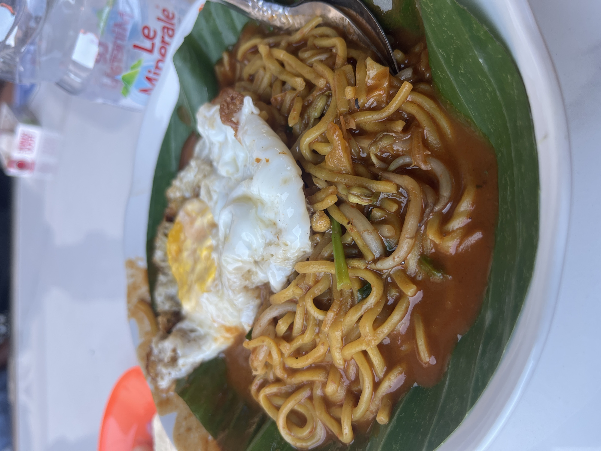 A close-up, eye-level shot shows a plate of Indonesian noodles served on a banana leaf, topped with a fried egg. The noodles are coated in a reddish-brown sauce and appear to be mixed with bean sprouts and green onions. A spoon rests on the side of the plate, partially submerged in the noodles. In the background, a plastic water bottle labeled "Le Minerale" and a red plastic bowl are visible, suggesting the meal is being enjoyed outdoors, possibly at a restaurant or food stall in Glameunasah Baro, Indonesia. The lighting is bright and natural, casting subtle shadows.