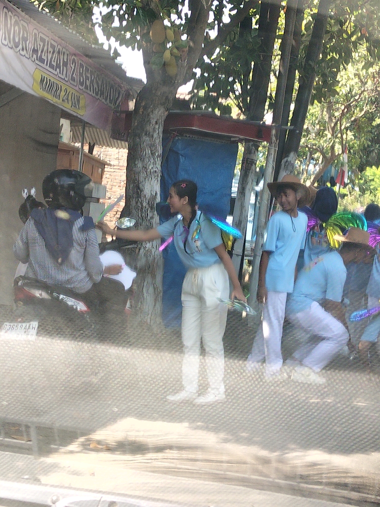 The image depicts an outdoor scene, likely in Kediri City, Indonesia, characterized by a bright, sunny atmosphere. In the foreground, a motorcycle with the license plate "G 3659 AAW" is parked. A person wearing a black helmet and a checkered jacket is seated on the motorcycle, holding what appears to be a white paper.
To the right of the motorcycle, a young person in a light blue uniform with iridescent fairy wings is interacting with the person on the motorcycle. This individual is holding a green, wing-shaped object. Another person, wearing a light blue t-shirt and a straw hat, is standing nearby, looking towards the right. Further in the background, other individuals are visible, some also wearing light blue shirts and hats. There are trees with fruit, possibly jackfruit, hanging from their branches, and a banner that reads "NOR AZIZAH 2 BERSINAR 24 JAM". The overall environment suggests a casual gathering or event taking place outdoors.