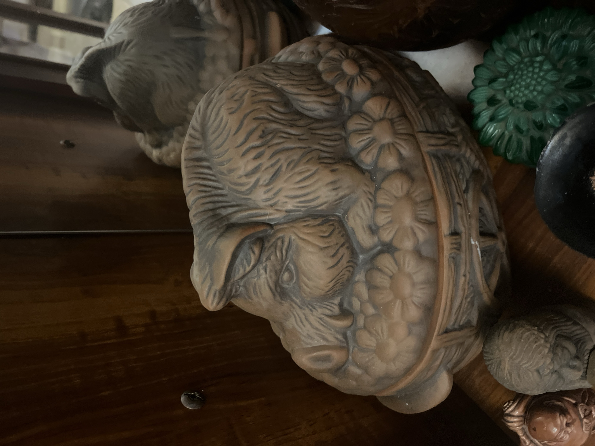 A terracotta lidded box, shaped like a sleeping boar resting on a basket of flowers, is displayed on a wooden shelf. The boar's face is detailed with closed eyes, textured fur, and floppy ears. The basket weave and flower details are also rendered in terracotta. Behind the boar box, another similar, but out-of-focus, object is visible. To the left of the boar box, part of a wooden cabinet with a visible knob is seen. In the foreground, to the right, a green, flower-shaped object and a dark, circular object are partially visible. The scene is illuminated by a soft light, casting subtle shadows. The location of Vladimirovka, Russia is not visible or inferable from the image.