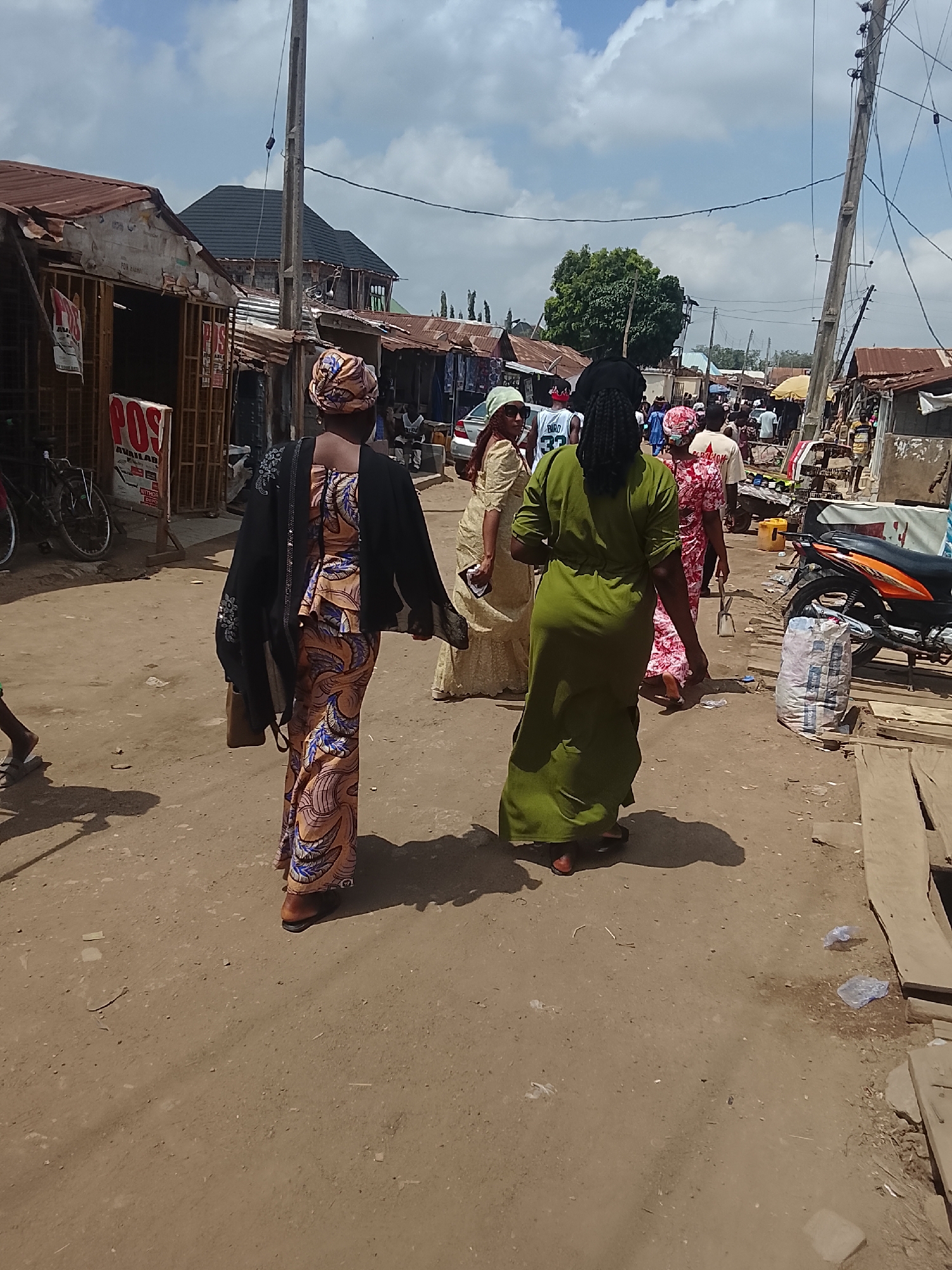 The image shows a street scene in Abuja, Nigeria, possibly a marketplace or neighborhood area with shops. The road is unpaved, dirt-covered with wooden planks providing pathways. The sky is blue with scattered clouds.
In the foreground, three women are walking away from the camera. The woman closest to the camera is wearing a colorful patterned dress and a black jacket. The next woman is wearing a green dress, and the third is wearing a light-colored dress.
Other people can be seen further down the street, including some wearing patterned clothing. A motorcycle is parked on the right side of the street with a bag next to it. On the left are shops with corrugated metal roofs. Electrical wires are strung overhead on poles.