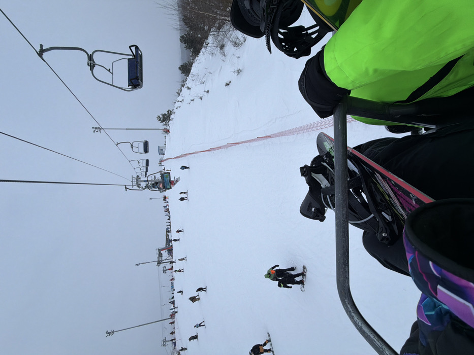 From a chairlift's perspective, a snowy ski slope descends. Several skiers and snowboarders are spread across the slope. A line of chairlift towers and chairs stretches into the distance under a cloudy sky. A snowboarder's boots and snowboard rest on the chairlift. A person wearing a neon green jacket and black gloves is also riding the chairlift.