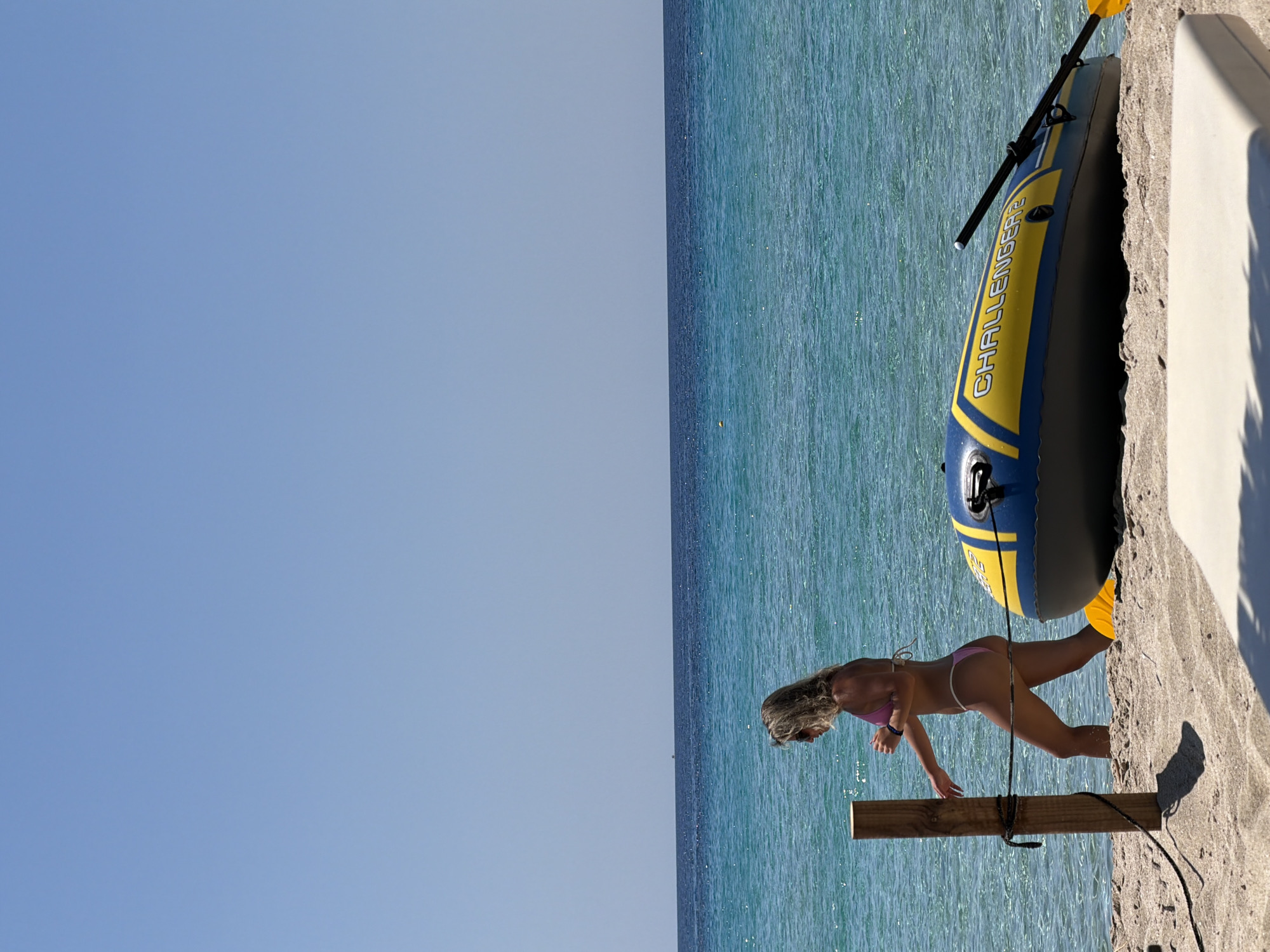 A woman in a pink bikini walks along a sandy beach next to the turquoise water of the ocean. A blue and yellow inflatable boat with the word "CHALLENGER" written on its side is partially submerged in the water. A wooden post with rope tied to it stands on the beach. The sky is clear and blue. The scene appears to be in Greece.