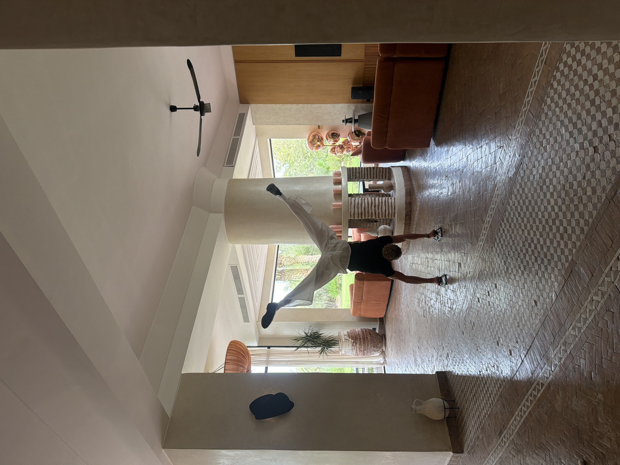 In a bright, spacious living area with high ceilings and large windows overlooking a lush green landscape, a person is performing a handstand. The individual, wearing a black t-shirt and loose white pants, is balanced on their hands with their legs spread wide in a V-shape. The floor is tiled with a mosaic pattern of terracotta and white ceramic. Orange sofas are positioned in the background, and a circular, multi-tiered fireplace with candles sits in the center of the room. A large, textured pot with a plant stands near a pillar on the left. A ceiling fan is visible overhead. The location appears to be Al Ouidane, Morocco, suggested by the architectural style and the surrounding vegetation.