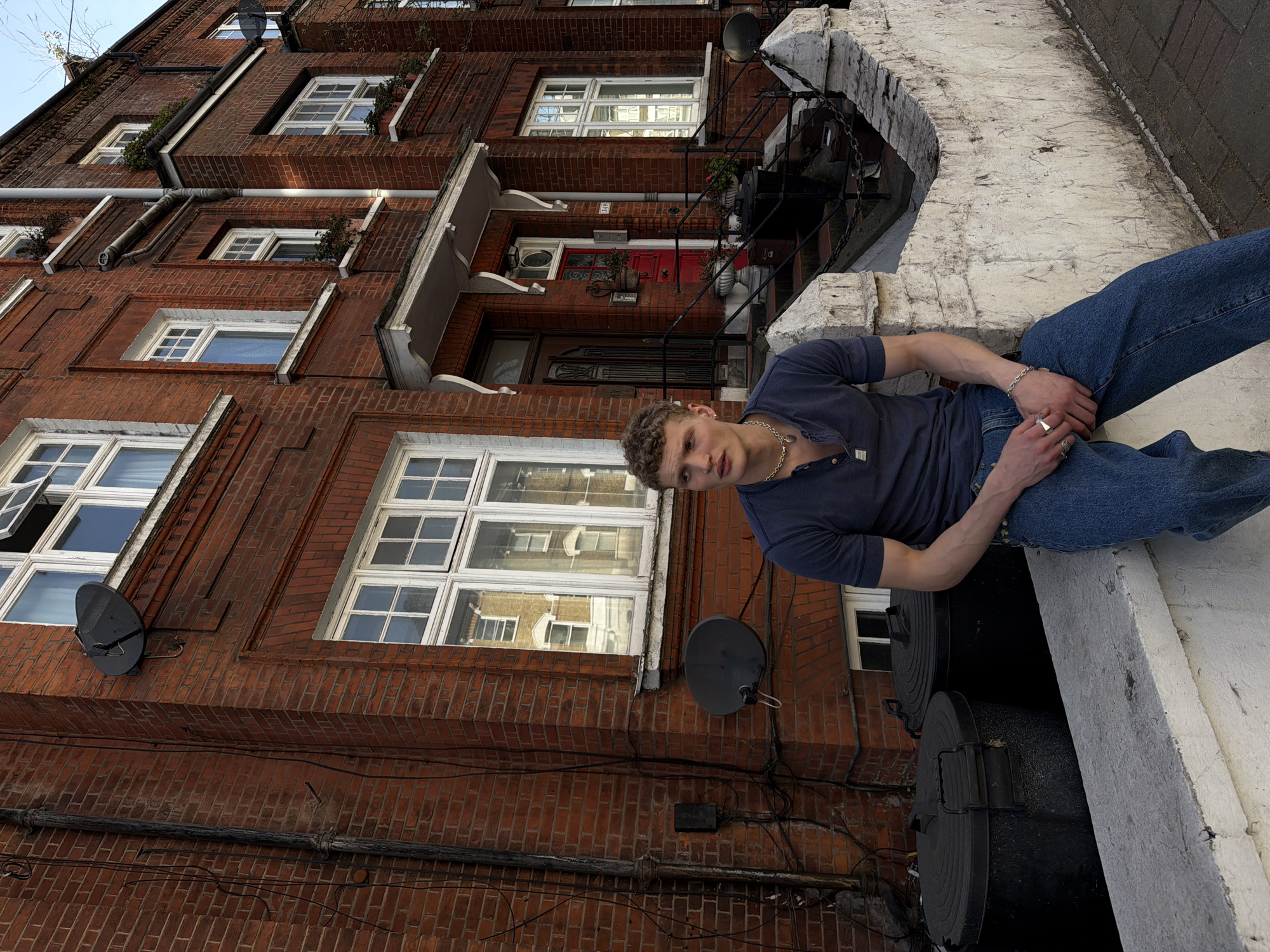 The photograph depicts a fair-skinned young man with short, curly blond hair, wearing a dark blue collared short-sleeved shirt, blue jeans, and a silver chain. He is sitting on a white ledge, with his hands resting in his lap, looking towards the camera. Below him are two large, black cylindrical containers. Behind him is a multi-story brick building with multiple windows and a red door. The building appears to be located in London, United Kingdom. A satellite dish is affixed to the building's facade.