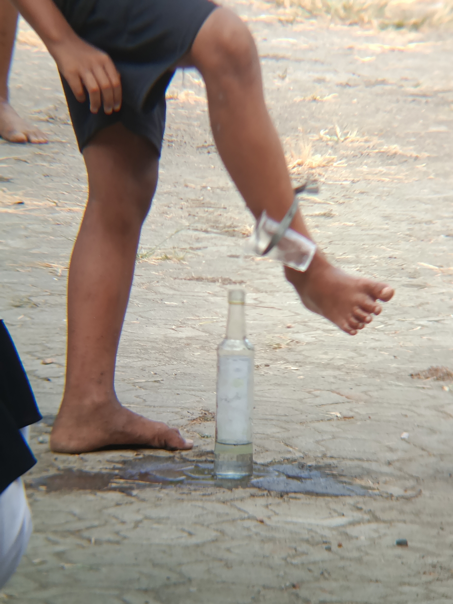 In Sarwadadi, Indonesia, a person's bare feet are visible on a dirt and gravel surface. A bottle of liquid stands upright on the ground, and a broken glass bottle is attached to the person's ankle. The person is wearing dark shorts.