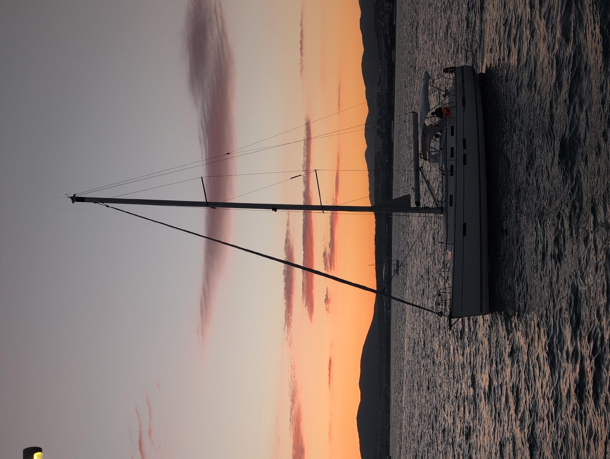 A wide shot captures a sailboat on the water during dusk, with the city of Gelendzhik, Russia, visible on the horizon. The sky displays a gradient of colors, from warm orange near the horizon to a soft pink and then a muted gray higher up, with wispy clouds scattered throughout. The sailboat, positioned in the center of the frame, is silhouetted against the vibrant sky. It has a tall mast and several lines running from it. The hull of the boat is white and appears to be moving through calm water. The water itself reflects the colors of the sky, with darker tones dominating the foreground. In the distance, mountains form a dark silhouette against the horizon, and a strip of land with buildings, presumably Gelendzhik, can be seen nestled between the water and the mountains. There are a few other, smaller boats visible further out on the water, also silhouetted. A single street lamp with a yellow light is visible in the upper left corner of the frame.