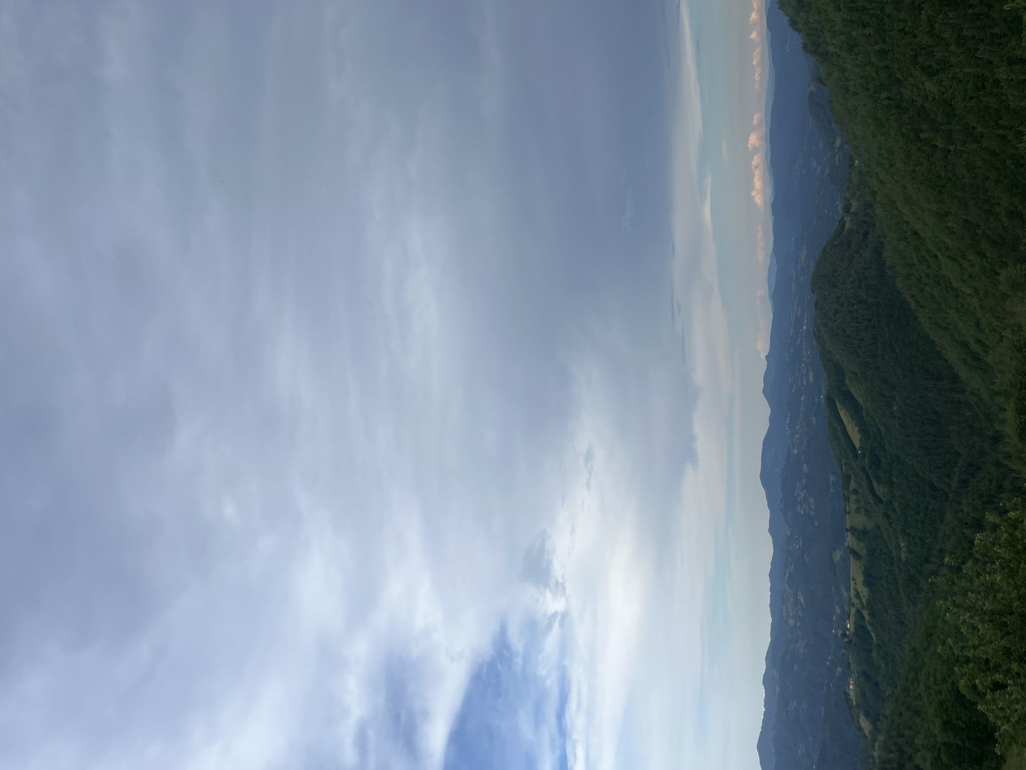 The image depicts a panoramic view of a hilly, forested landscape under a cloudy sky. The foreground features lush green trees, with the terrain receding into a series of rolling hills covered in dense foliage. In the mid-ground, scattered houses and agricultural fields are visible on the slopes, indicating human habitation and land use. The background is dominated by layers of distant mountains, fading into a hazy blue silhouette against the sky. The sky itself is a canvas of dramatic clouds, with patches of lighter blue sky peeking through, suggesting a dynamic weather system. The overall scene conveys a sense of natural beauty and rural tranquility. There are no people or specific objects that would indicate a particular location within Startsevo, Bulgaria.