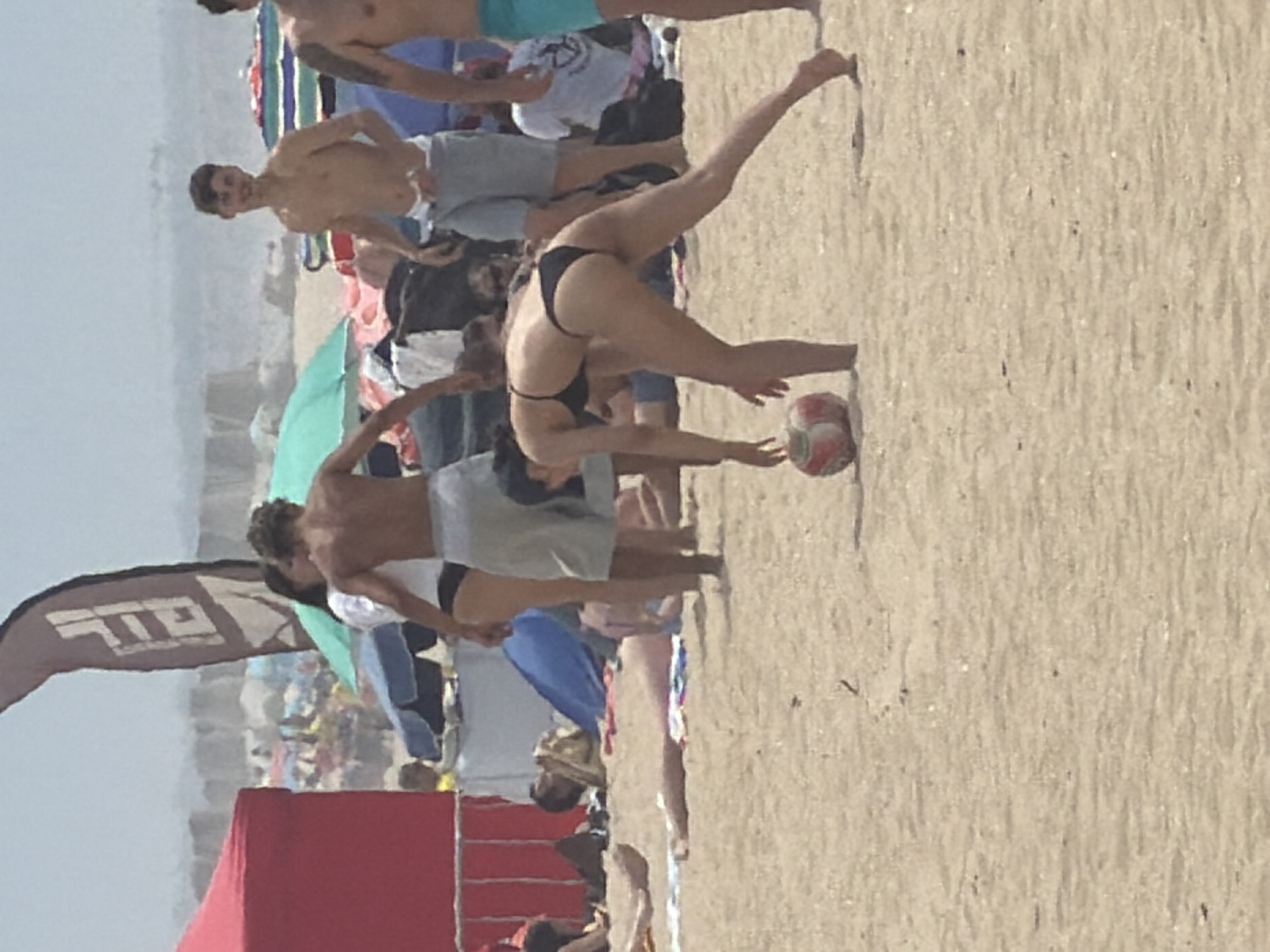 The image captures a lively scene on a sandy beach, likely in Espinho, Portugal, judging by the context provided. In the foreground, a woman in a black bikini is playing with a red and white ball. Her body is bent, suggesting she is about to kick or has just kicked the ball. Behind her, a man with a muscular build, wearing light-colored shorts, stands with his arms by his sides. Further back, other beachgoers are visible, some sitting on towels or under colorful umbrellas. A large red tent is prominent on the left side of the image, and a tall, dark banner with white lettering is visible in the background, partially obscured by people and beach equipment. The overall atmosphere appears to be a sunny, active day at the beach, with people enjoying leisure activities.