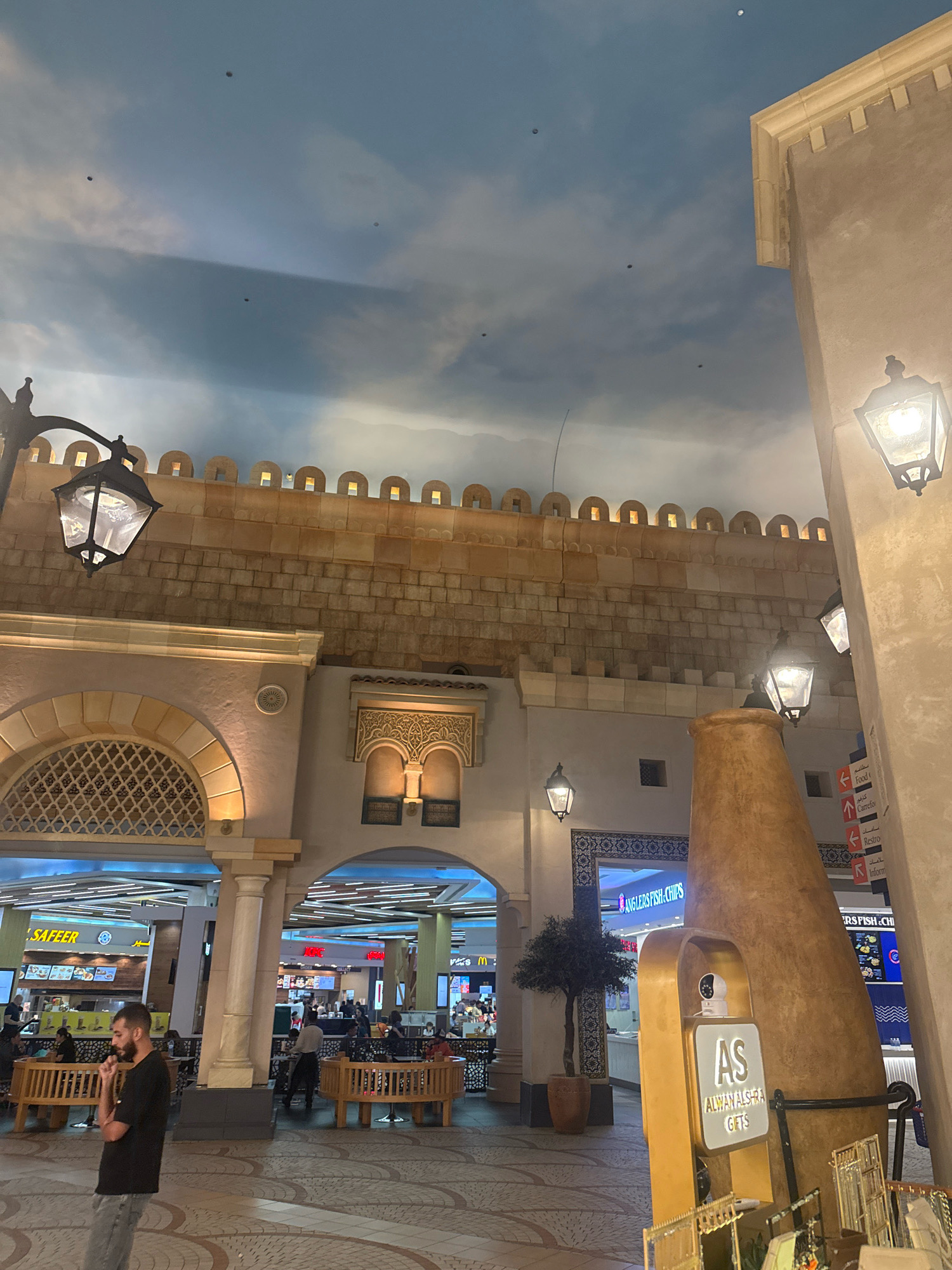 The image depicts an indoor scene designed to resemble a traditional Arabian marketplace, with a vaulted ceiling painted to mimic a cloudy sky. Architectural elements include sandstone-colored brickwork, arched doorways with decorative latticework, and ornate windows with intricate carvings. Several ornate street lamps are visible, some illuminated. In the foreground, a man wearing a black t-shirt and grey pants stands facing left, appearing to be talking on his phone. To the right, a large, conical, terracotta-colored structure is prominent, with a sign that reads "AS ALWAN ALSHRA GIFTS" and a security camera mounted on top. In the background, a food court area is visible with various dining establishments, including "SAFEER," "MCDONALD'S," and "ANGLERS FISH & CHIPS." People are seated at tables and walking through the food court. The overall impression is a modern commercial space designed with an immersive, historical theme, likely located in a shopping mall in Dubai, United Arab Emirates, given the architectural style and signage in English and Arabic.