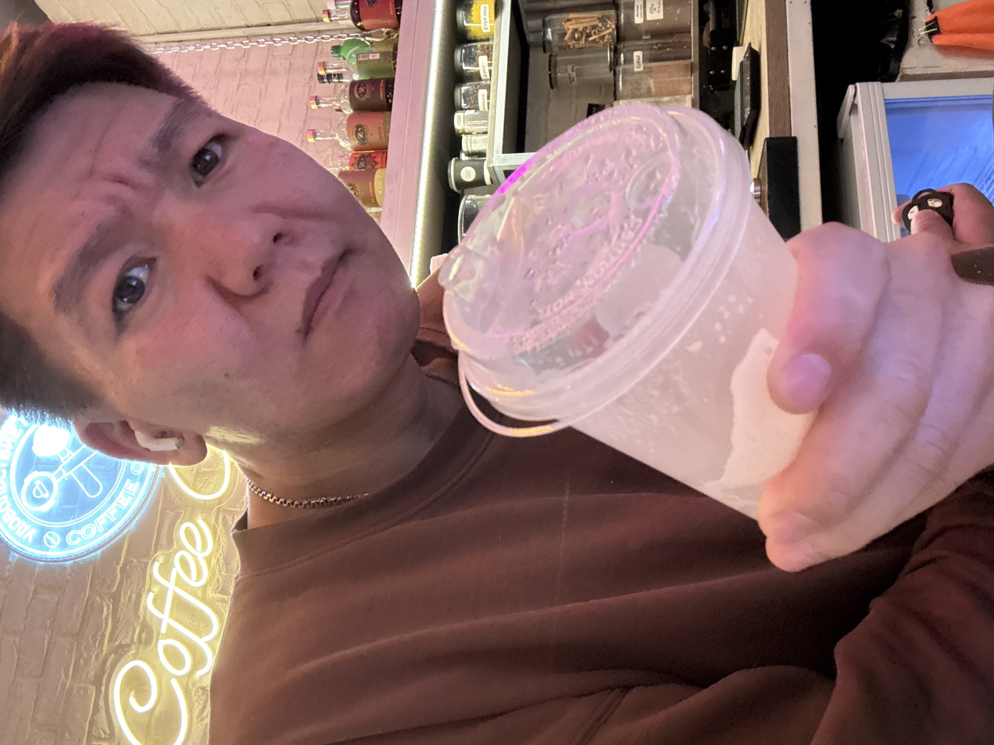 This close-up, angled selfie features an Asian male in what appears to be a cafe or bar setting in Moscow, Russia. The man, with short dark hair and reddish highlights, looks directly at the camera with a slightly scrunched, contemplative expression. He is wearing a dark brown long-sleeved top, a thin silver chain, and a white earbud in his left ear.
He holds a clear plastic cup with a lid in his right hand, containing a light-colored beverage with ice. The lid has embossed text, including what appears to be "HOT". The background is filled with elements typical of a coffee shop or bar. On the left, a yellow neon sign vertically spells "Coffee". Below it, a circular blue neon sign displays Cyrillic text, including "COFFEE," "ВИДЕО" (Video), and "РОБОТА" (Work/Robot), along with a key and coffee cup icon. Behind the man, shelves are stocked with various bottles and clear containers holding ingredients like spices or coffee beans. On the far right, another hand is visible holding a small black electronic device or key fob, positioned near a white refrigerator or cabinet. The scene is lit by warm indoor lighting, suggesting it is evening.