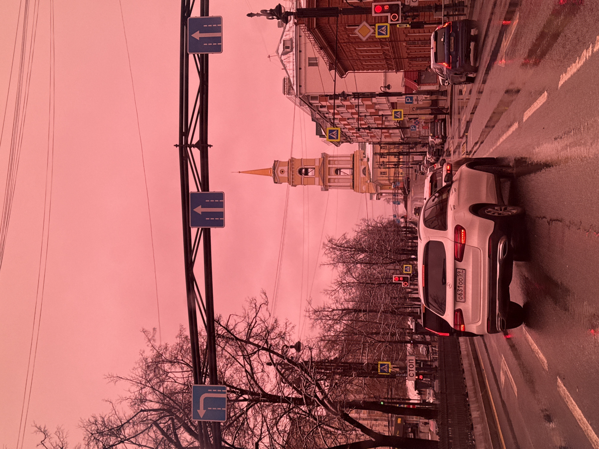 The image, which is rotated 90 degrees counter-clockwise, depicts a city street in Perm, Russia, under an unusually vivid pinkish-red sky. The wet asphalt road reflects the sky and streetlights, indicating recent rain.
In the foreground, a tall metal pole on the left supports multiple blue rectangular traffic signs with white arrows, two indicating "straight ahead" and one pointing for a "right turn." Bare-branched trees line the left side of the street, their silhouettes visible against the colored sky.
On the wet road, a white SUV is prominently featured in the mid-ground, its rear visible to the viewer. Its Russian license plate, "063 100 59," is clearly legible. Several other vehicles are seen further down the street, along with painted road markings for lanes and crosswalks.
Along the right side of the street, a row of multi-story buildings with varied architectural styles forms the backdrop. A distinctive light-colored building with a prominent yellow, spire-topped tower stands out in the mid-distance. Street furniture includes lampposts, a red traffic light, and various signs such as pedestrian crossing indicators, 'P' signs for parking, and a white rectangular sign with the Cyrillic word "СТОП" (STOP). No people are distinctly visible. The scene portrays an active urban environment under dramatic or uniquely colored atmospheric conditions.
