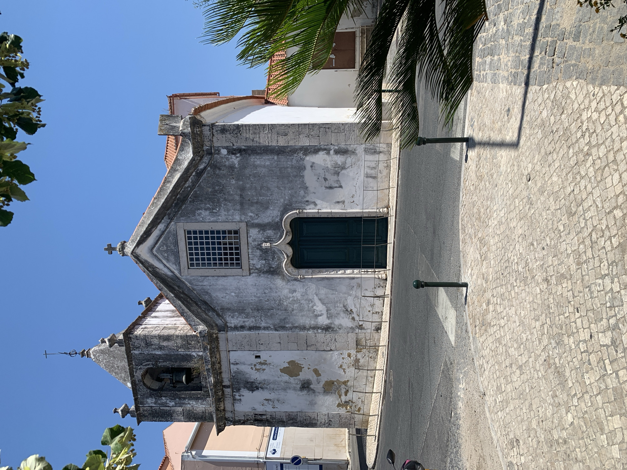 This image depicts the front facade of a weathered, historic church under a clear blue sky in Odivelas, Portugal. The church, constructed of stone with a whitewashed exterior that shows significant signs of age and peeling, features a prominent bell tower on the left side. The tower is topped with a pyramidal roof and a small cross. The main body of the church has a pitched tile roof and a large, arched window with a grid pattern above an ornate, dark green double door. To the right of the main structure, a white section of the church with a terracotta-tiled roof is visible.
In the foreground, the street meets a cobblestone sidewalk. The street itself is paved with asphalt, and a pedestrian crossing is marked. Two dark green bollards are positioned on the sidewalk, one casting a long shadow across the cobblestones. A palm tree with fronds obscures part of the right side of the image. In the background, to the left of the church, a light pink building is partially visible, with some signage indicating it might be a school. The scene appears to be a quiet, sunny day, with no people or vehicles actively engaged in the scene, aside from a glimpse of a motorcycle on the far left.