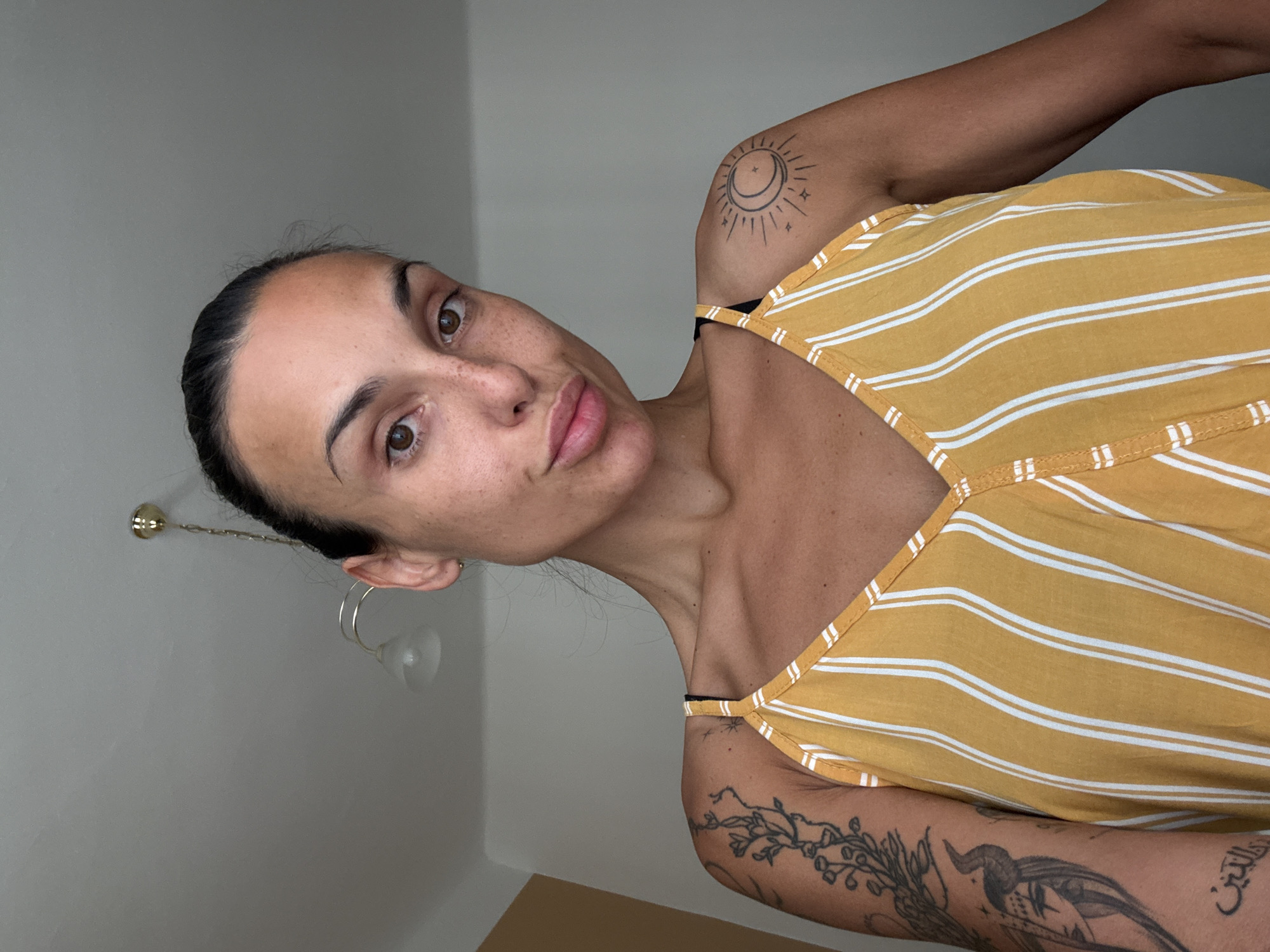 A young woman is looking at the camera from a low angle. She has her hair pulled back and is wearing a yellow and white striped top. She has tattoos on her arms, including a sun and moon design on her shoulder. The background appears to be an indoor setting with a white wall and ceiling. There are two light fixtures visible on the ceiling. The lighting suggests it is daytime. There is no indication of the location being Prague, Czechia.
