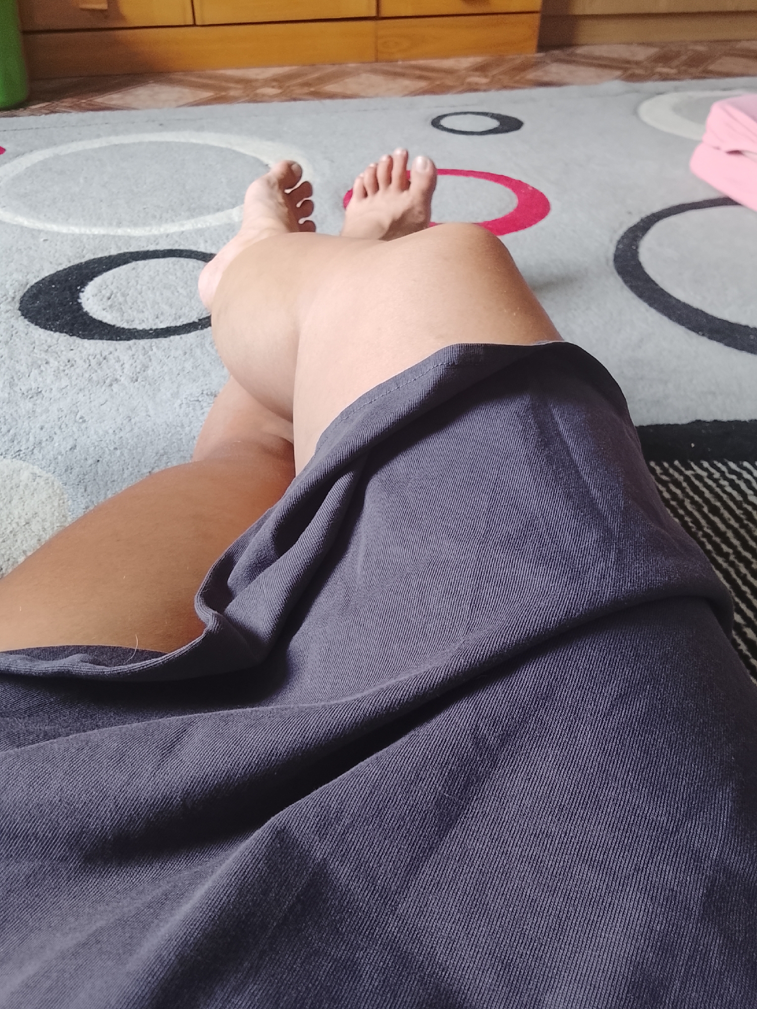 The media file shows a close-up view of a person's legs and feet resting on a patterned carpet. The person is wearing a dark grey skirt that drapes over their legs. Their feet are bare and positioned towards the top of the frame, with toes clearly visible. The carpet has a design of concentric circles in various colors, including grey, black, white, and pink. In the background, there is a hint of wooden furniture and possibly a portion of a pink object, which could be a cushion or a piece of furniture. The lighting suggests it is daytime, and the indoor setting appears to be a living space or bedroom. There are no visible text elements in the media file. The scene depicts a moment of relaxation or rest.