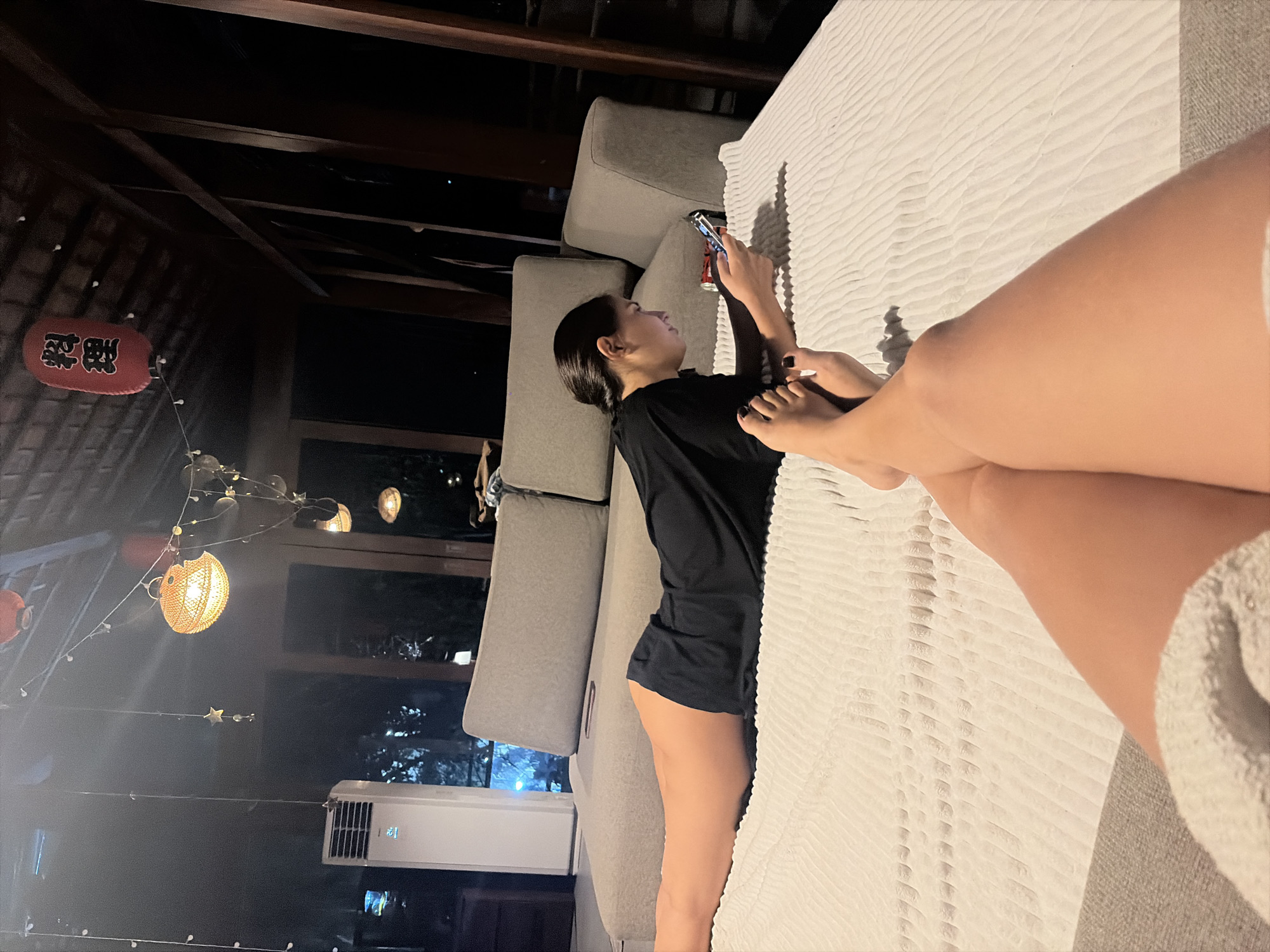 The image depicts an indoor scene at night, likely on a balcony or veranda given the exposed wooden beams and roof structure. A young woman with dark hair pulled back is lying on a light-colored textured surface, possibly a mattress or large cushion, and is looking at and interacting with her smartphone. She is wearing a black t-shirt and dark shorts. In the foreground, another person's legs and feet are visible, also resting on the same surface, with the feet crossed and painted toenails. In the background, a grey sectional sofa is visible, along with an air conditioning unit. The environment is dimly lit by string lights and several hanging lanterns, one of which is red with Japanese characters. The scene appears to be located in Kediri, Indonesia.
