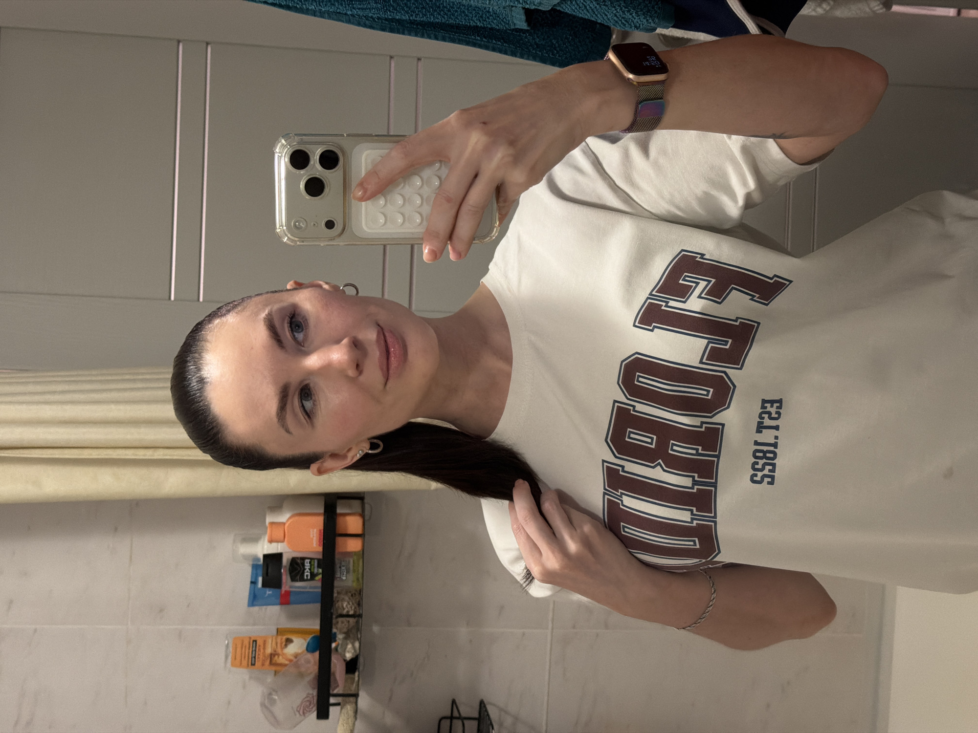 A woman is taking a selfie in a bathroom with white tiled walls. A shower curtain and bathroom toiletries are visible. The woman has dark hair pulled back in a ponytail and is wearing a white t-shirt with "FLORIDA EST. 1855" printed on it. She is holding a smartphone in her right hand with a smartwatch visible on her wrist. A bracelet is visible on her left wrist. The location is not discernible from the image. Therefore, it cannot be confirmed whether the location is in Perm, Russia.