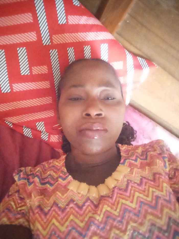 The image displays a close-up, eye-level perspective of an adult female subject positioned indoors, appearing to be lying down. The female has dark-toned skin, dark eyes, and dark, curly hair visible on the right side of her head. Her lips are slightly parted and appear glossy. She wears small, gold-colored hoop earrings. Her garment is a patterned top featuring a multicolor zigzag (chevron) design in hues of red, orange, yellow, blue, and white. The neckline is adorned with distinct, light yellow/tan, rounded rectangular elements. A dark-colored layer is visible beneath the patterned top's collar. Behind her head is a red pillowcase with a repeating geometric pattern of white and black striped rectangles and diagonal lines. A wooden surface is visible in the upper right background, and a solid dark red fabric object is present in the lower left. The scene is located in Unguwan Rimi, Nigeria.
