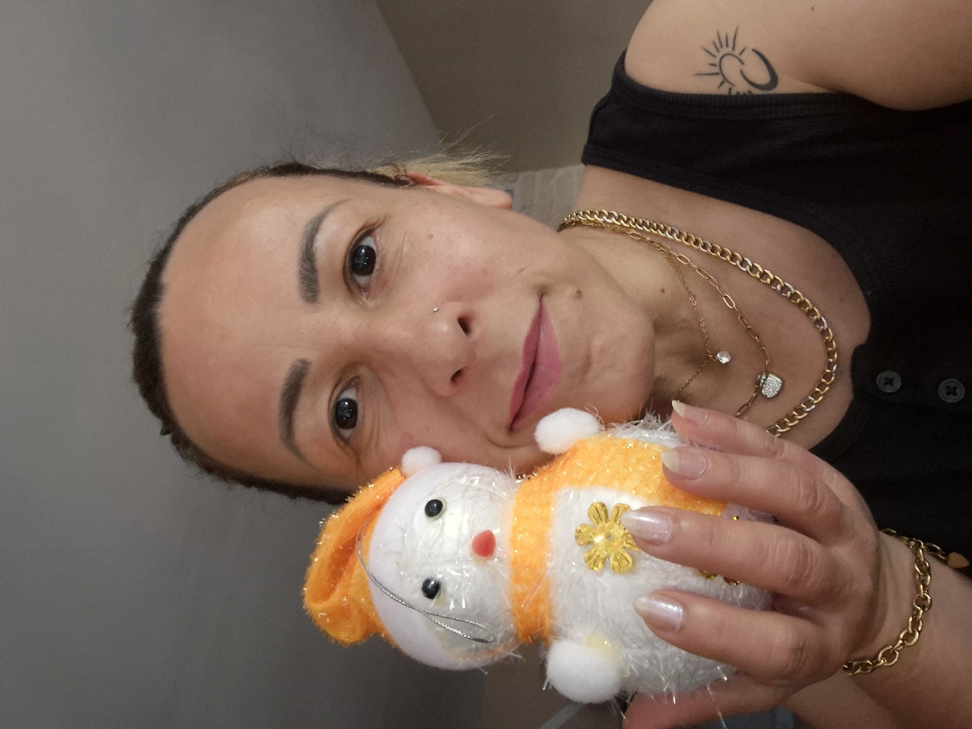 A woman poses indoors, holding a stuffed animal. She has light brown skin, dark eyebrows, a pierced nose, and reddish lips. Her hair is dark at the roots and blonde at the ends. She wears a black tank top and multiple gold necklaces and a bracelet. On her upper left arm is a tattoo of a sun and moon. The stuffed animal is white with orange trim and has a gold flower attached. The location appears to be in a private residence, possibly Unknown, Unknown.