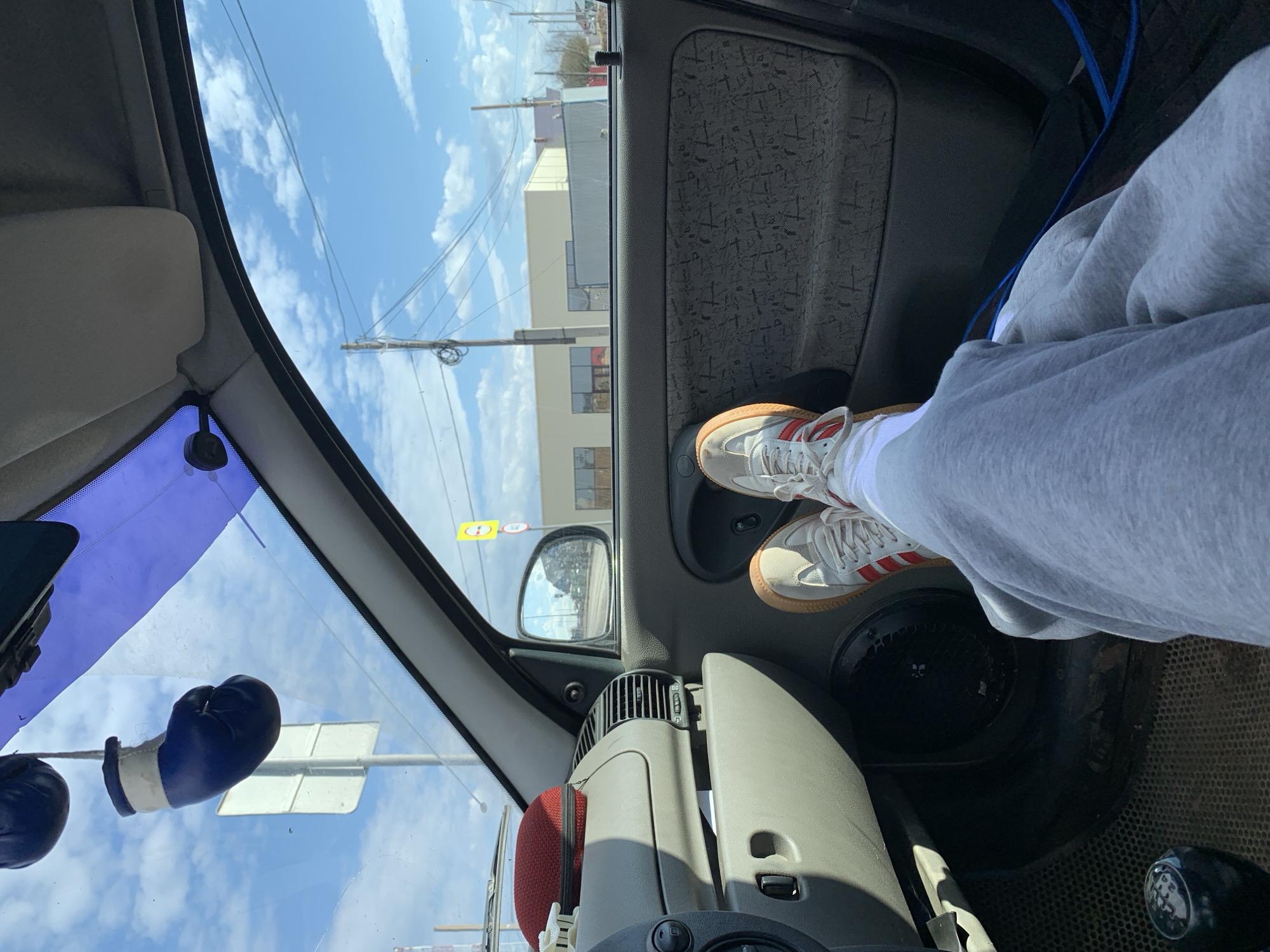 The image captures an interior view from a car, showing a person's feet propped up against the door on the passenger side. The person is wearing grey sweatpants, white socks, and white sneakers with red Adidas stripes and a gum sole.
The car interior features a dark grey door panel with a textured fabric insert, an air vent, and a dashboard section. A gear shift knob, indicating manual transmission with numbers 1 through 5 and R, is visible on the lower right. On the dashboard, a red cushion or accessory is present. Miniature blue boxing gloves hang from the rearview mirror, which reflects parts of the exterior.
Through the windshield and side window, a clear blue sky with scattered white clouds is visible. The exterior scene shows utility poles with numerous wires, and several buildings in the background, appearing to be commercial or industrial structures. A yellow circular road sign with a black border and a black symbol (possibly a 'T' or other traffic indicator) is seen outside the window. The overall setting suggests a daytime scene in an urban or semi-urban area. The location context is Гаревая, Russia.