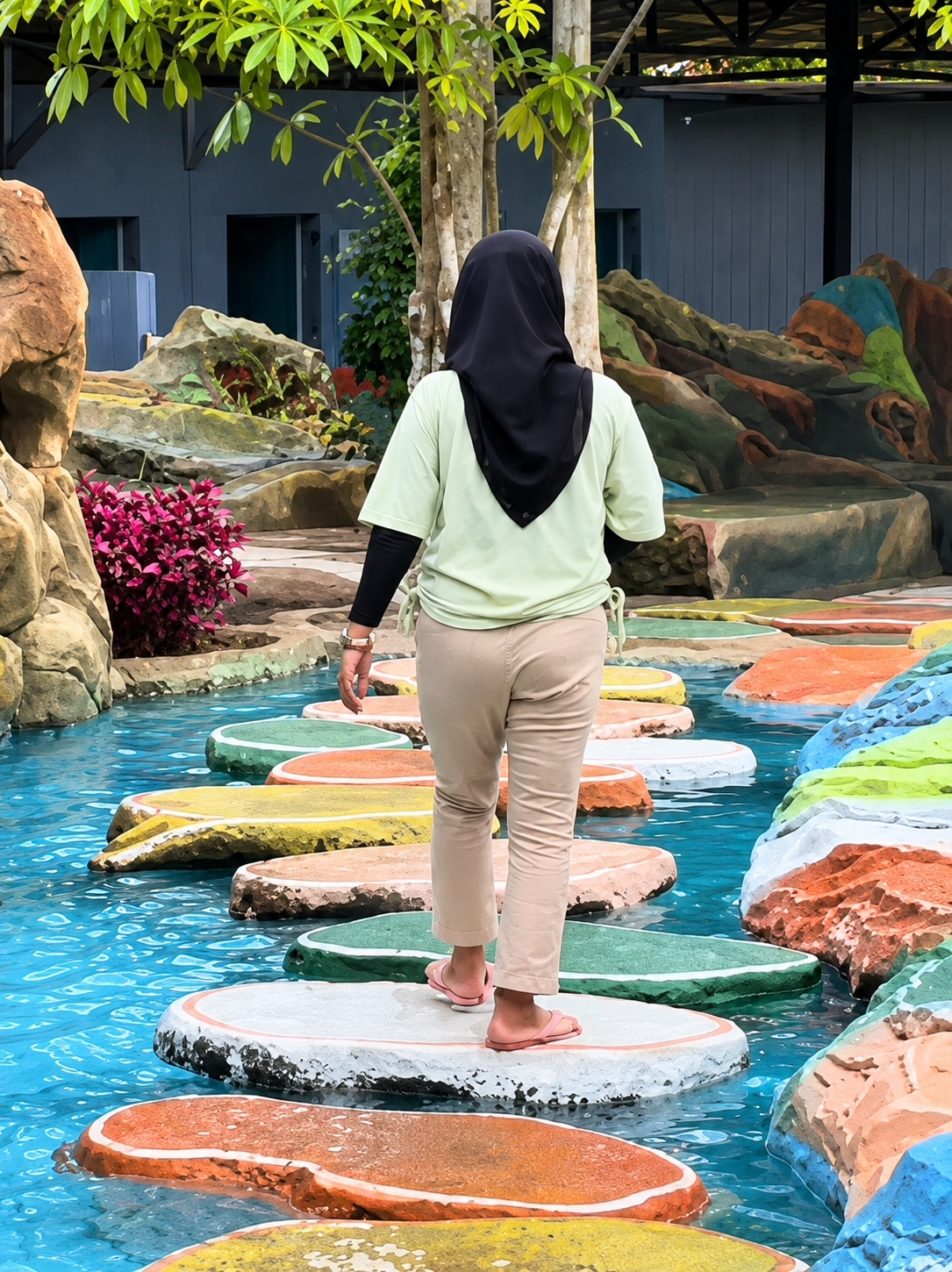A person wearing a black hijab, a light green long-sleeved shirt, and beige pants walks across a path of colorful, irregularly shaped stepping stones in a shallow blue pool of water. The stepping stones are painted in various bright colors, including orange, yellow, green, and white, with some showing hints of other colors like red and blue. The person is facing away from the camera and is mid-stride, with their right foot on a white stepping stone and their left foot about to land on another. In the background, there are large, man-made rock structures painted in vibrant, earthy tones, as well as lush green foliage and trees. The overall setting appears to be a recreational or decorative area, possibly a theme park or a landscaped garden within a resort. The image suggests a warm, outdoor environment. The person's attire indicates adherence to modest dress. The water is clear, allowing the colors of the stepping stones to be seen beneath the surface.