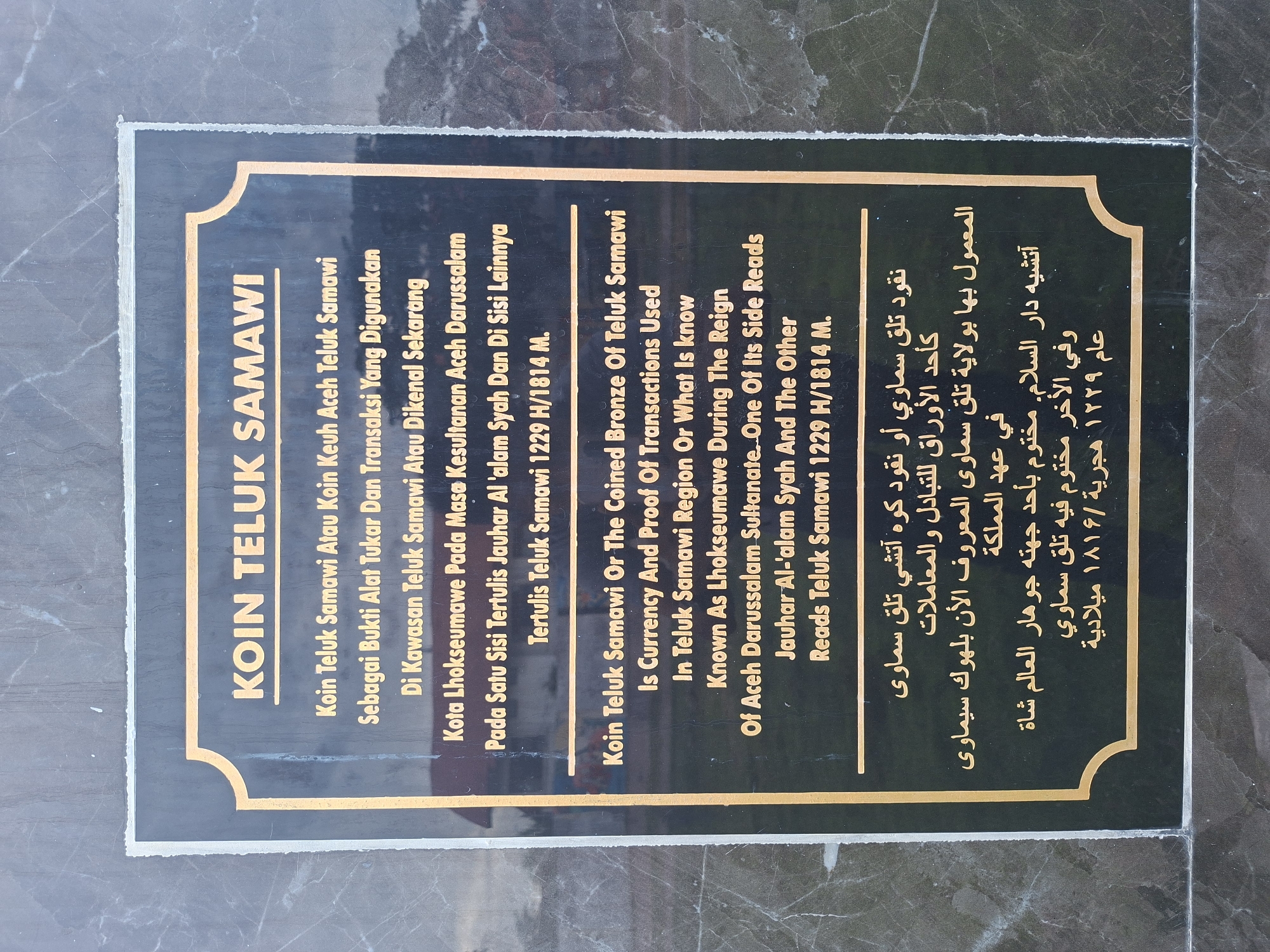 The image displays a rectangular, dark-colored informational plaque with gilded text and a golden border, featuring decorative curved corners. The plaque is set against a dark, mottled surface, possibly marble or granite, suggesting a museum or historical site setting. Good lighting illuminates the golden lettering.
The plaque provides information about the "KOIN TELUK SAMAWI" (Teluk Samawi Coin) in three languages: Indonesian, English, and Arabic.
The text explains that the Teluk Samawi Coin, also referred to as the "Coined Bronze of Teluk Samawi" or "Koin Keuh Aceh Teluk Samawi," served as a form of currency and proof of transactions. It was utilized in the Teluk Samawi region, which is presently known as Lhokseumawe City, during the period of the Aceh Darussalam Sultanate. The inscription further details that one side of the coin bears the name "Jauhar Al-'alam Syah," while the other side reads "Teluk Samawi." The date of issuance or circulation is given as 1229 H (Hijri) / 1814 M (Gregorian) in the Indonesian and English sections. Notably, the Arabic section records the Gregorian year as 1816 AD, presenting a minor discrepancy in the date compared to the other two languages.