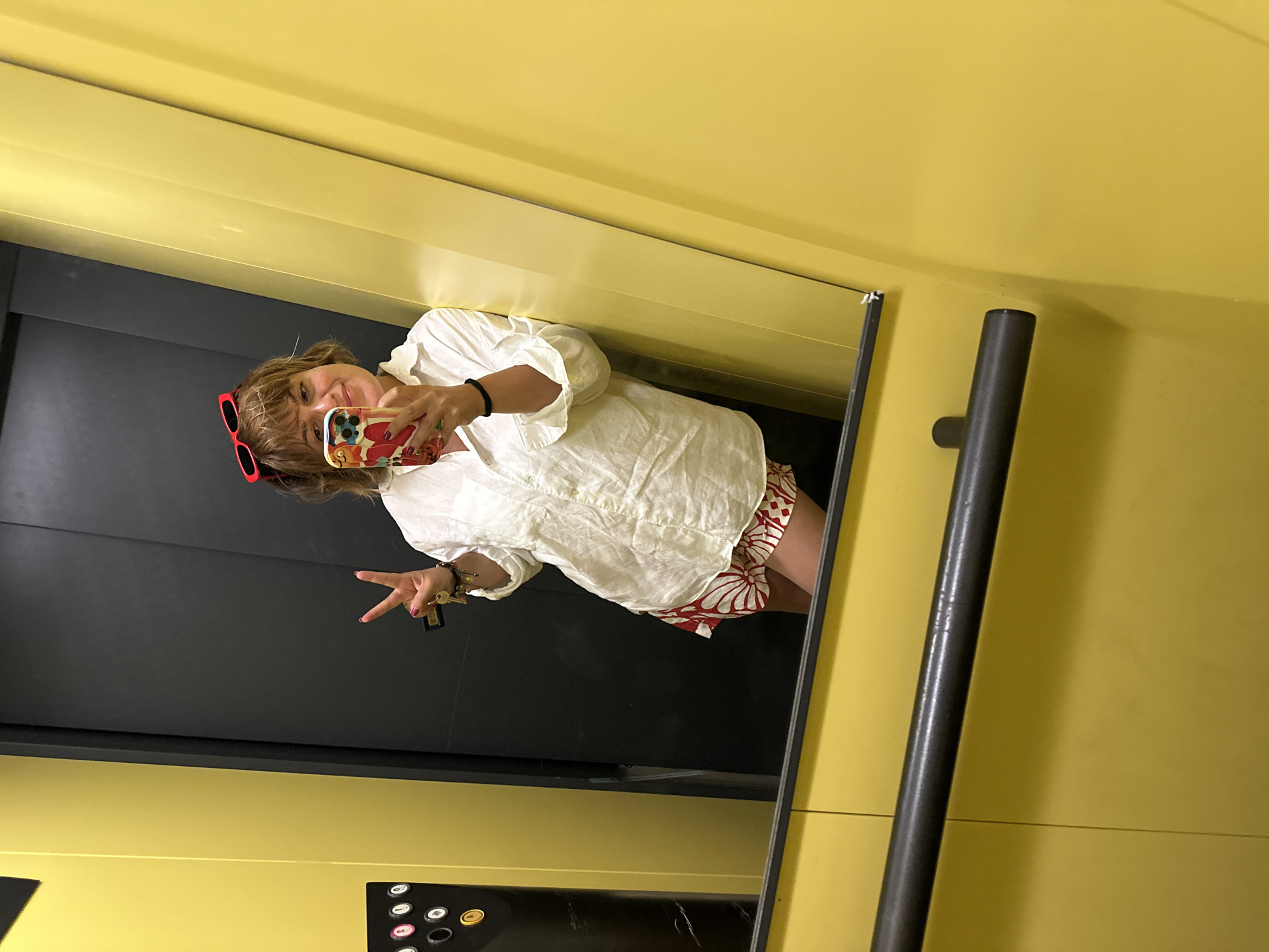 A woman with short, light brown hair, wearing red sunglasses on her head, a white oversized button-down shirt, and red and white patterned shorts, stands in an elevator. She holds a smartphone in her right hand, taking a selfie. Her left hand is raised, forming a peace sign. She wears several bracelets on her left wrist, and a black band on her right wrist. The elevator walls are bright yellow, and the doors are dark grey. A metal handrail runs across the lower part of the image. The scene appears to be in an elevator in Pineto, Italy.