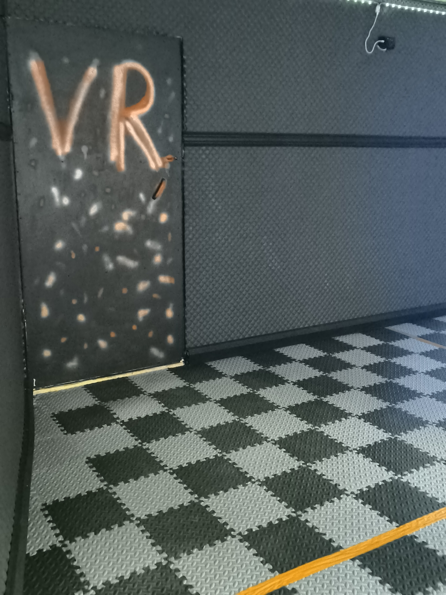 The image displays an empty indoor space, likely a dedicated area for virtual reality (VR) experiences, located in Trastenik, Bulgaria. The room is characterized by dark tones and functional design elements.
On the left side of the frame, a dark, vertical partition or wall segment is prominent. It features the letters "VR" spray-painted in a bold orange color, clearly indicating the room's purpose. This partition also shows areas with white and orange speckles, possibly from paint application or wear. Its base meets the floor with a narrow, light-colored trim.
The main wall stretching across the background is covered in a dark gray, textured material, which appears to be padded or sound-dampening, common in VR environments. A thin black horizontal strip runs across this wall at approximately half its height. Along the top edge of the wall, a white LED light strip provides illumination. A small, dark electronic device, likely a VR tracking sensor or camera, is mounted on the wall near the top right corner, with a white cable extending downwards from it.
The floor is entirely covered with interlocking foam mats, arranged in a distinctive checkered pattern of dark gray/black and lighter gray squares. An orange tape or cord runs diagonally across the lower right portion of the floor, potentially marking a boundary or a safety zone for VR play.
The room is devoid of people, and no activity is currently in progress. The overall impression is that of a prepared, functional space designed for immersive virtual reality sessions.