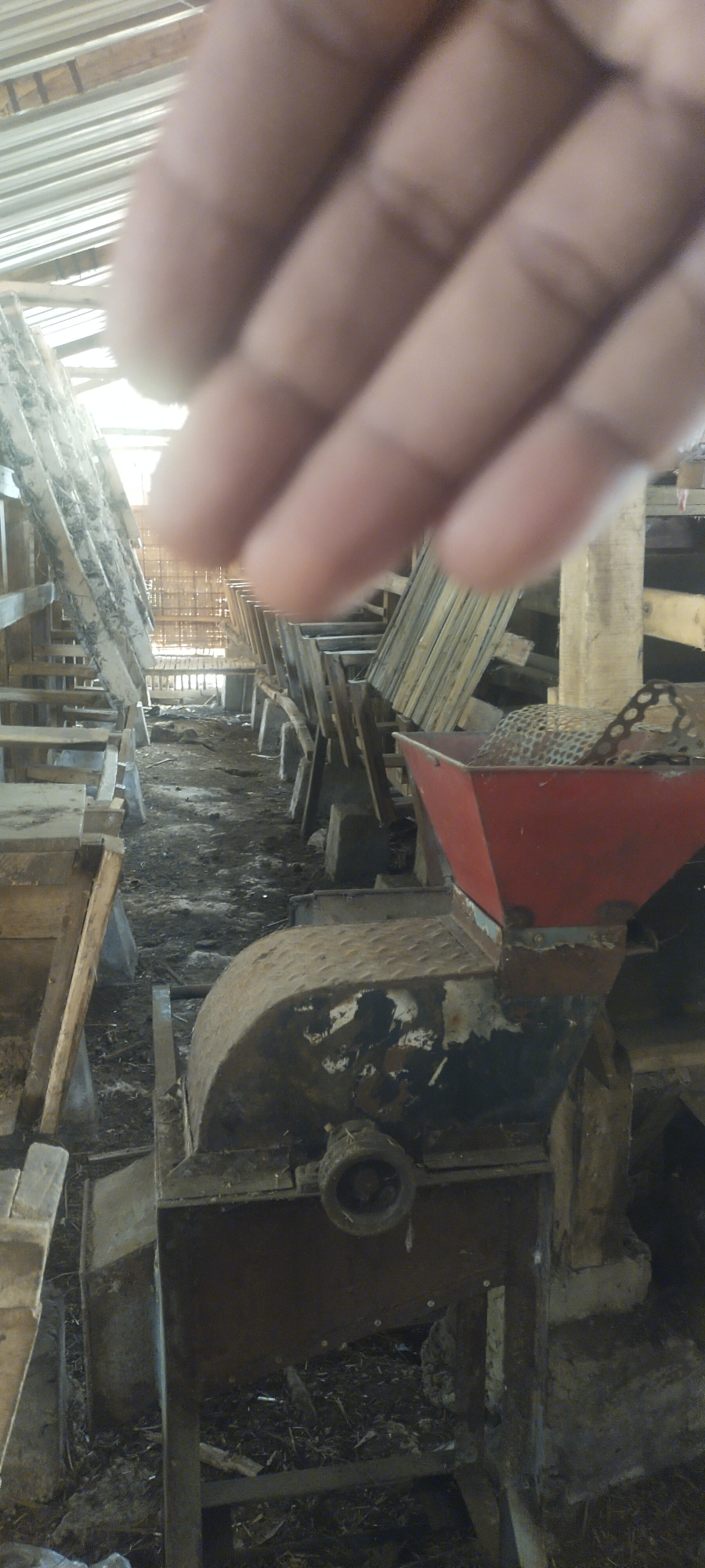 The image shows a close-up view of a hand obscuring the upper portion of the frame. The lower portion of the image reveals a rustic interior scene, possibly a shed or agricultural facility in the city of Pasirhuni, Indonesia. In the foreground, a worn, metal machine with a rusted, semi-circular covering and a red hopper is visible. The machine appears to be industrial or agricultural equipment, possibly a grinder or processing unit. The ground is covered with dirt, debris, and what looks like straw or dried organic matter. To the left and right of the central machine, wooden structures with angled slats are visible, suggesting they might be troughs, feeders, or storage compartments. The overall environment appears functional and utilitarian, with evidence of agricultural activity. The lighting suggests an indoor setting with some natural light filtering in from above.