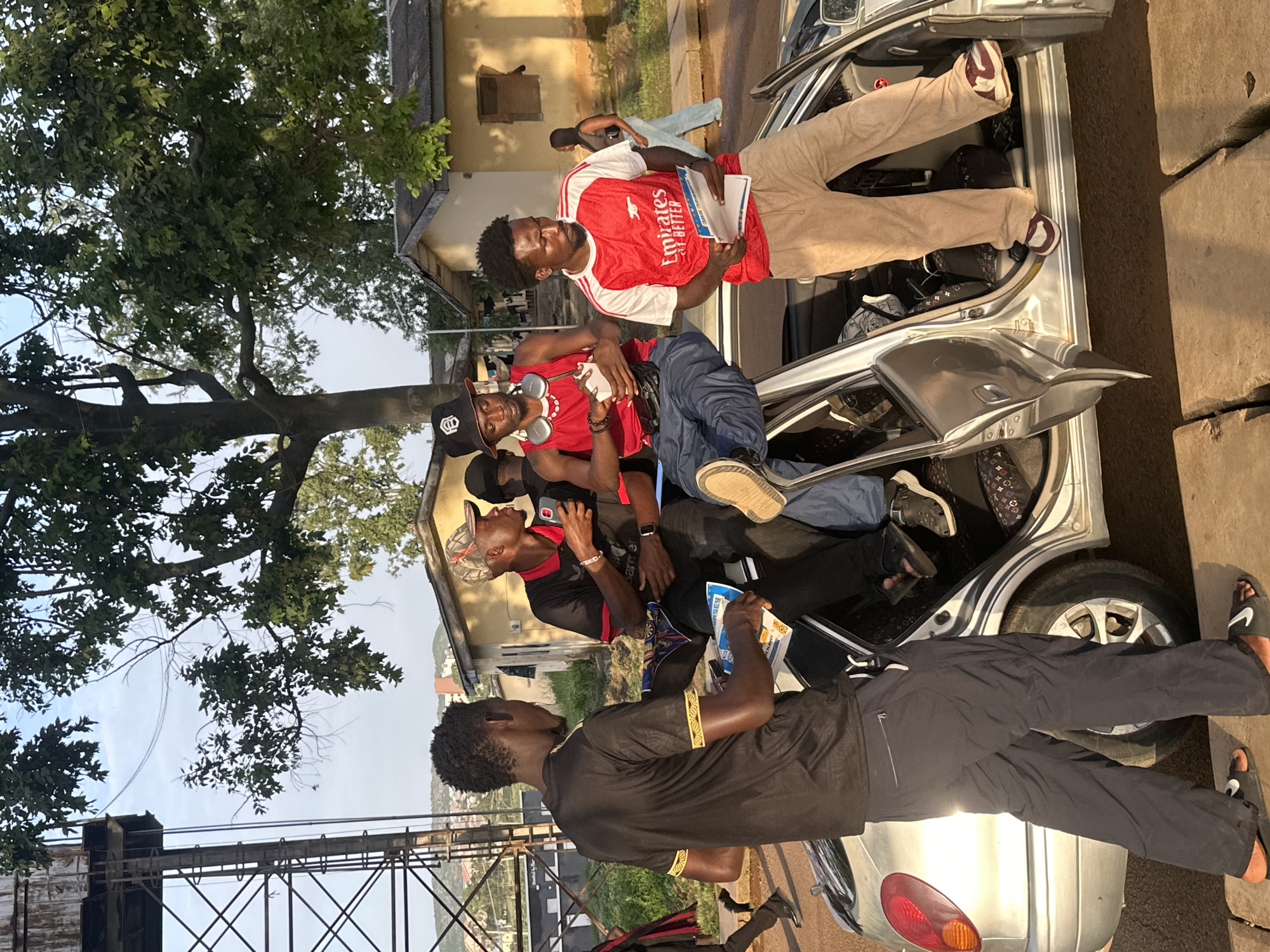 The image depicts a group of young men in what appears to be an urban or semi-urban setting in Rafin Par, Nigeria. Several individuals are positioned inside and around a silver car, which is open, revealing the interior and trunk.
In the foreground, a young man wearing a black shirt with gold trim is partially visible, with his legs stretched out. He is wearing dark pants and dark sandals with a prominent white logo. Behind him, another man in a black shirt and a camouflage baseball cap is looking upwards and holding a phone. To his left, a man in a red and white Arsenal jersey is sitting inside the car, holding papers. He has dark skin and a beard. Further inside the car, another individual in blue pants and a red shirt is visible. The back of the car also shows another person's legs wearing a light brown tracksuit and burgundy and white sneakers.
The scene suggests a casual gathering or perhaps a moment of pause during an activity. The lighting indicates it is daytime, likely afternoon, with bright sunlight casting shadows. The background shows buildings, some appearing residential, and lush green trees, indicating a possibly warm and humid climate typical of Nigeria. A metal structure, possibly part of a bridge or overpass, is visible in the distance.
The Arsenal jersey has text on it, including "Emirates" and "FLY BETTER." The papers held by the man in the Arsenal jersey also appear to have some text, but it is not clearly legible. The overall mood seems relaxed and informal.