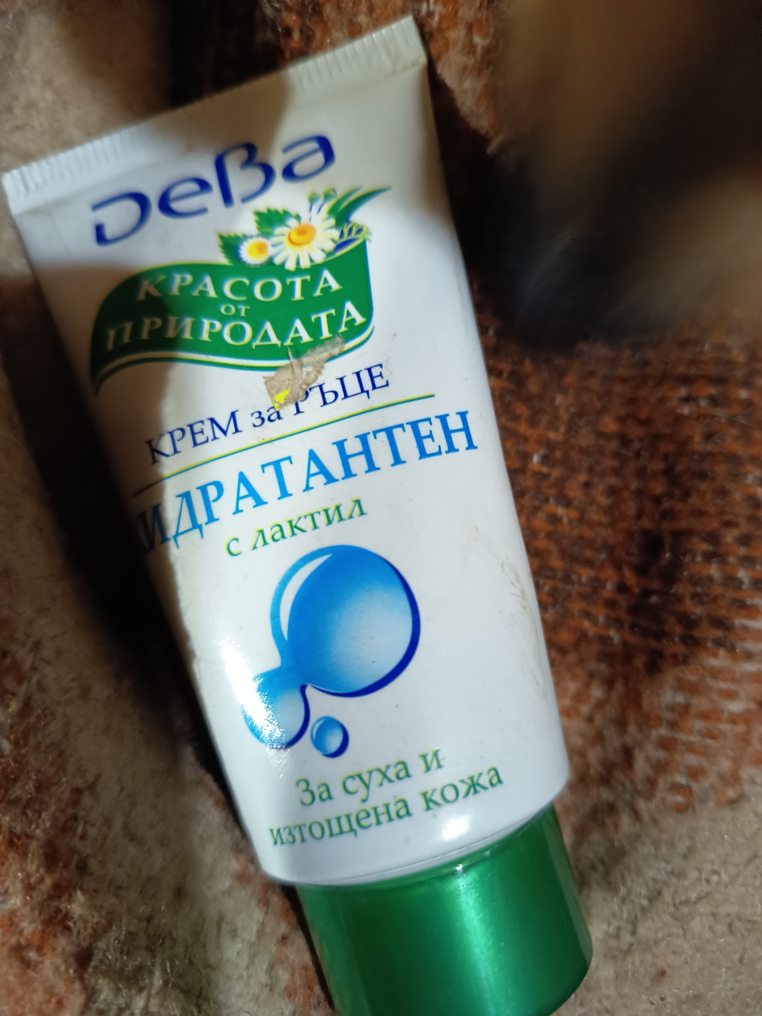 Here is a description of the image:
The image shows a tube of "Deba" hand cream lying on a textured surface, possibly fabric. The tube is predominantly white with green accents, featuring text in Bulgarian. The text includes phrases like "КРАСОТА ОТ ПРИРОДАТА" (Beauty of Nature), "КРЕМ за РЪЦЕ" (Hand Cream), "ИДРАТАНТЕН" (Hydrating), and "За суха и изтощена кожа" (For dry and exhausted skin). A graphic with a chamomile flower and blue water droplets are also visible. Part of the image is out of focus, particularly in the upper right area.