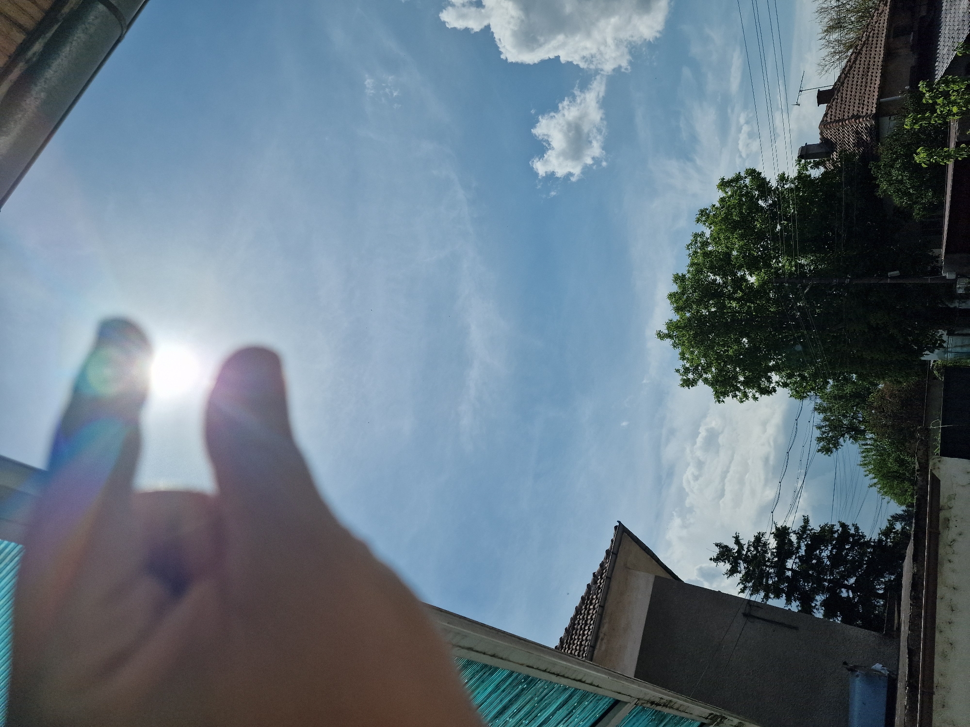 The image is a low-angle shot of a sunny sky with scattered clouds, taken from what appears to be an enclosed or semi-enclosed area. In the foreground, a blurred hand is positioned to frame the sun, creating a heart shape with the fingers. The surrounding environment includes rooftops and trees, typical of a residential or suburban area. The architectural styles visible suggest a location within a Central European or Balkan context. Given the specified location of Byala Slatina, Bulgaria, the scene is likely within a residential area of that city. Overhead power lines run parallel to the roofline in the distance. The angle implies the photographer is either looking up from a low vantage point, or the camera is angled upwards.