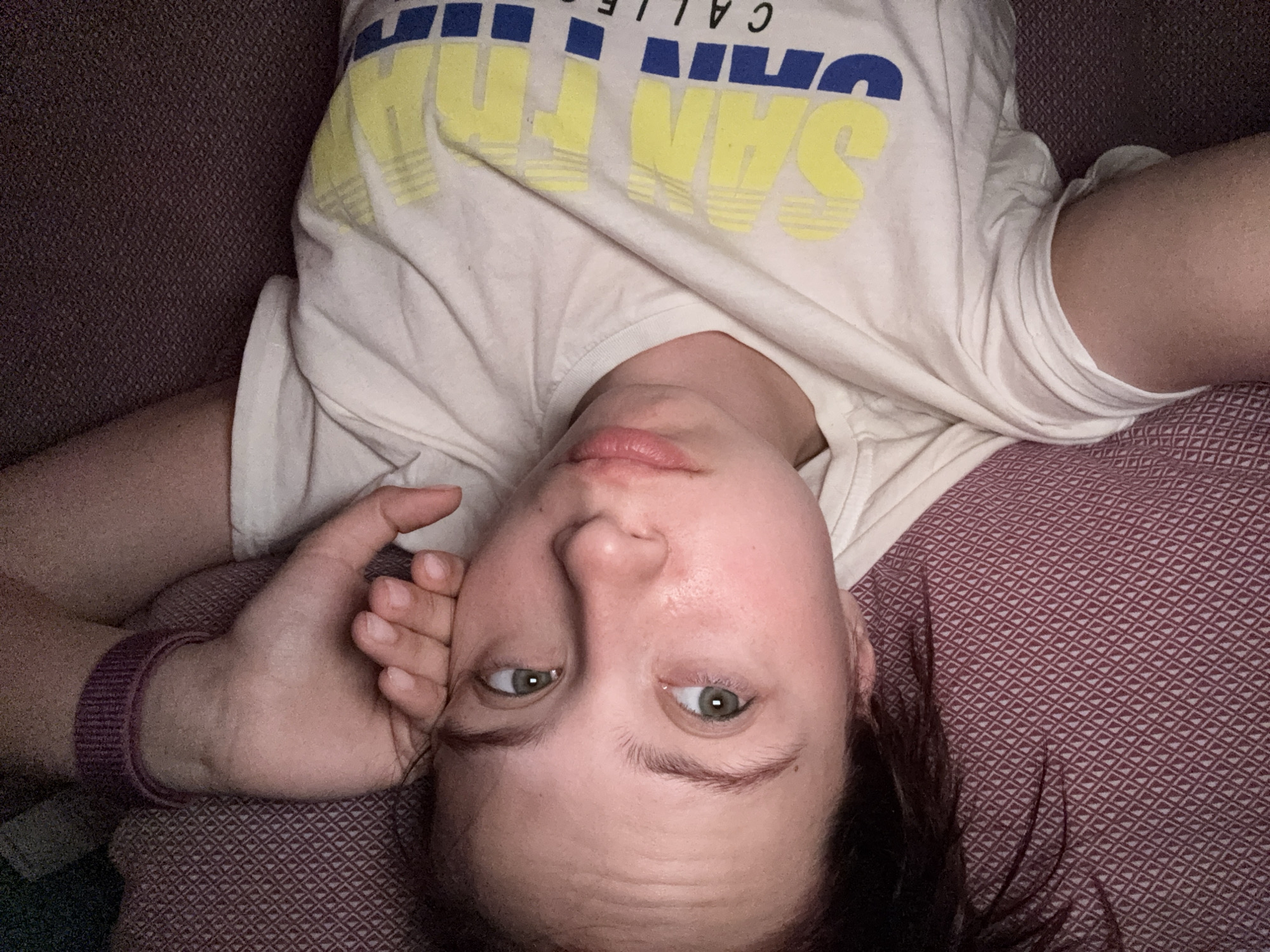 A person is lying upside down on a patterned surface, possibly a couch or bed. They are wearing a white t-shirt with the words "SAN CALLE" printed on it in yellow and blue. Their left hand is near their face, and they are looking towards the camera. The patterned surface is red/maroon with a repeating geometric design. The location is unknown and cannot be deduced from the image.