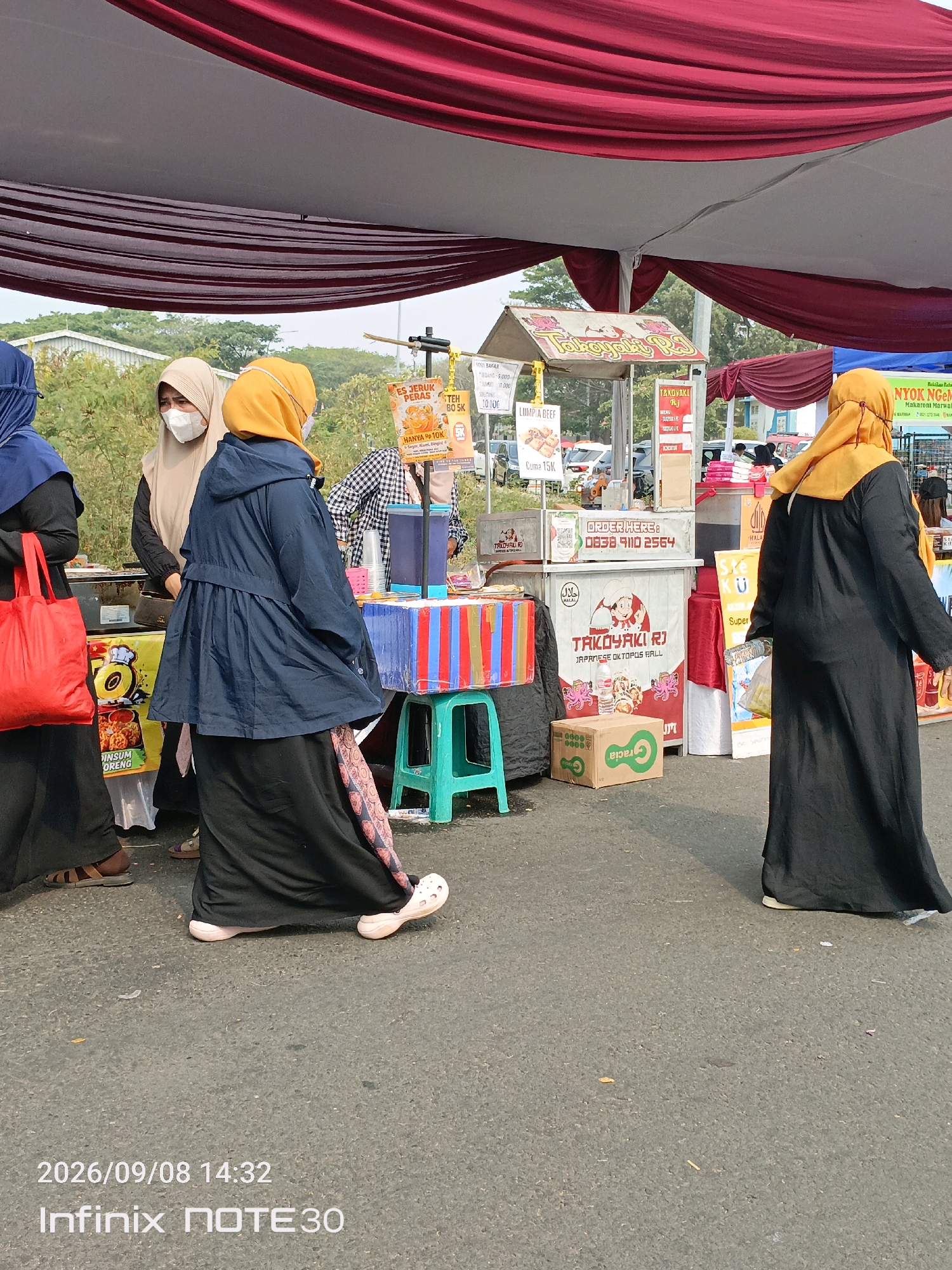 The image shows an outdoor scene, likely a market or food stall area, under a large tent with burgundy and white fabric draped overhead. Several people are visible, predominantly women wearing Islamic attire, including hijabs. Some are interacting with food stalls, while others are walking.
One woman in the foreground on the left wears a blue hijab and a black dress, carrying a bright red tote bag. To her right, another woman wears a beige hijab and a white mask, looking towards a food stall. A third woman, centrally positioned, wears a navy blue hooded jacket over a black dress and a patterned skirt, with an orange hijab. She appears to be walking away from the viewer. Further back, another woman in an orange hijab stands at a food stall.
The food stalls feature colorful signage advertising various food items. One stall has a sign with "TAKOKOYAKI RJ" and "JAPANESE OCTOPUS BALL". Another sign advertises "ES JERUK PERAS TEH BOSK" (Fresh Orange Juice Tea) for 10K. There are also boxes and containers associated with food preparation and sales.
The ground is paved asphalt, with some small debris visible. In the background, there are trees and glimpses of vehicles, suggesting an urban or suburban setting. The overall atmosphere appears to be that of a bustling marketplace or event.
The timestamp at the bottom left corner reads "2026/09/08 14:32", and "Infinix NOTE30" is also visible, indicating the device used to capture the image. While the image itself doesn't explicitly state the location as East Jakarta, Indonesia, the context of outdoor markets and food stalls with the Indonesian language on the signs strongly suggests this region.