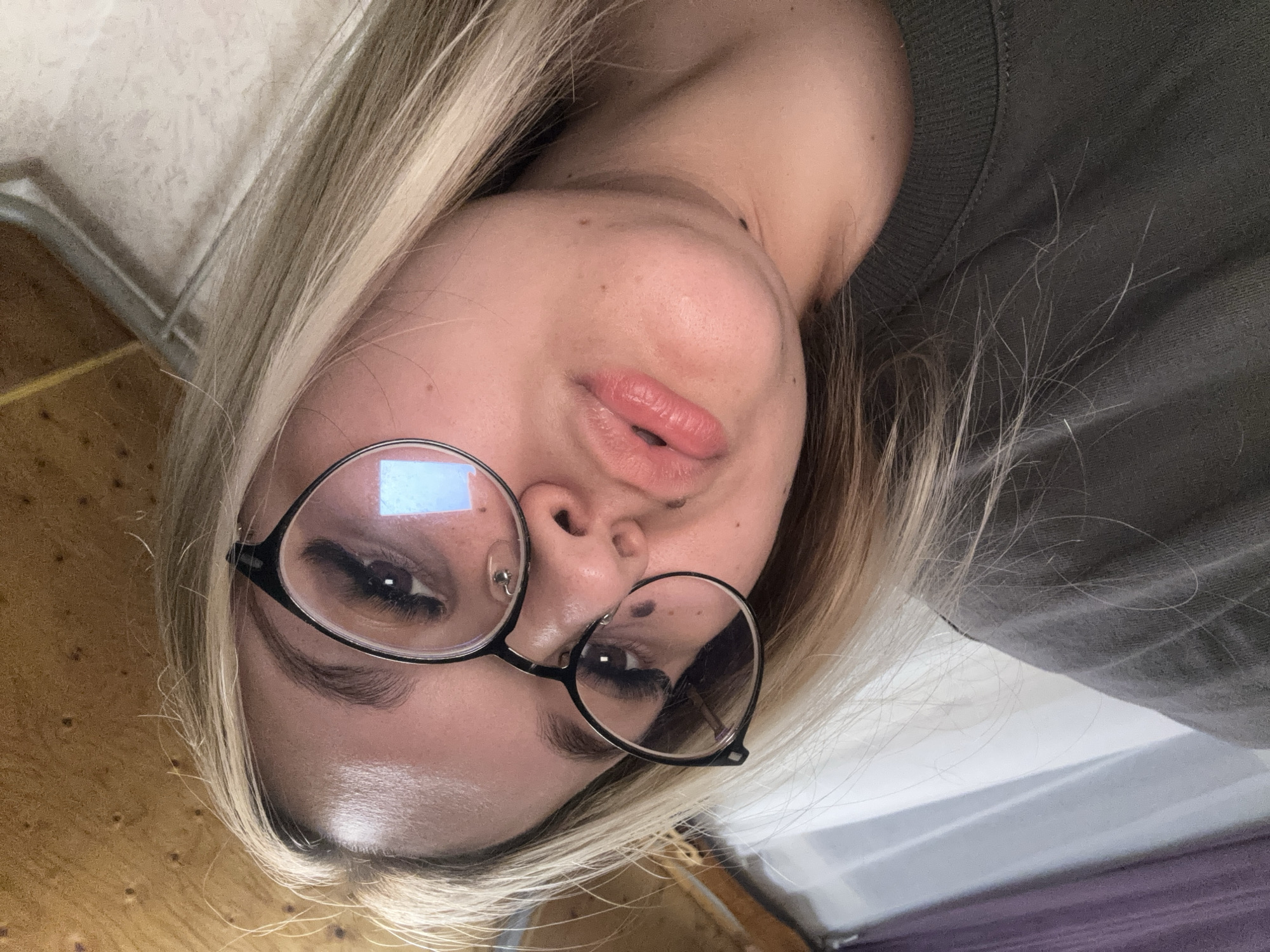 A rotated close-up selfie captures a young individual, likely female, with fair skin and long, light blonde hair. The subject is wearing prominent round, black-framed glasses. Through the glasses, their dark eyes and distinctly long eyelashes are visible, with a bright, rectangular light source reflected in the left lens. Their lips are slightly parted, showcasing a glossy, light pink finish. Several small moles or freckles are scattered across their cheeks and chin, and a subtle nose stud piercing is visible on the left nostril. Dark, well-defined eyebrows contrast with the blonde hair.
The person is wearing a dark, possibly olive green or grey, sweater with a ribbed collar partially visible. The setting is indoors, with a textured, light-colored surface (likely a wall or ceiling) in the upper left background and a wooden surface (possibly flooring or furniture) visible on the left and lower right sides. Another light-colored, possibly white, surface is in the lower right background. The lighting appears to be artificial and internal. The image's significant rotation suggests it was taken from a low angle, looking upwards, or while the subject was lying down. The expression is neutral and calm. No discernible text is visible in the image.