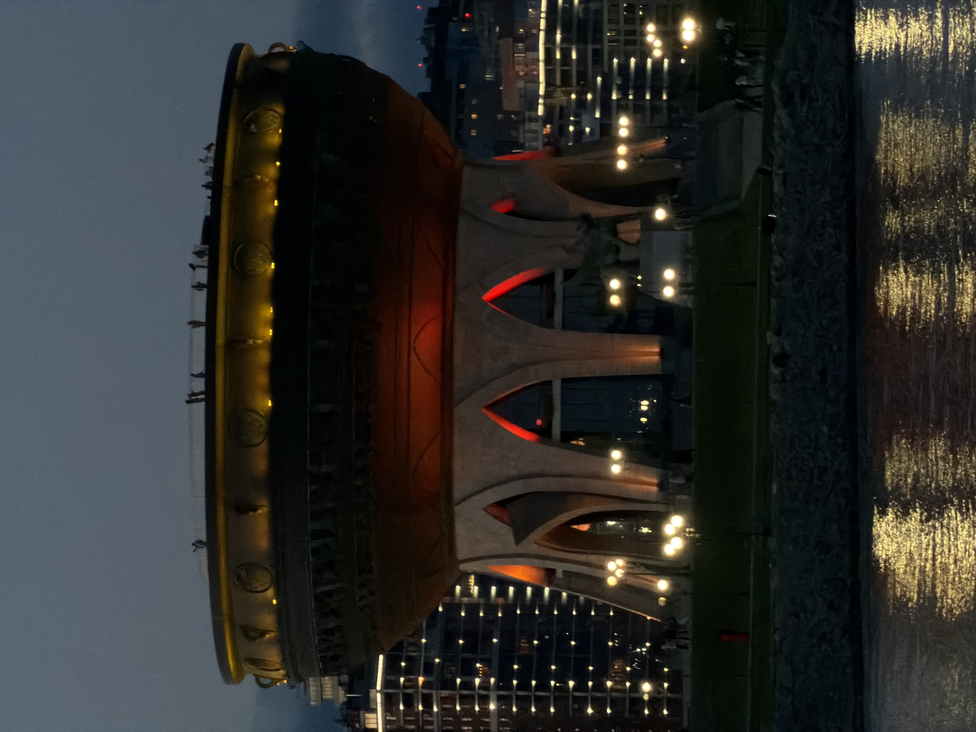 A nighttime, eye-level shot depicts the Kazan Family Center, located in Kazan, Russia. The structure is situated alongside a body of water, with reflections of city lights shimmering on its surface.
The building is partially illuminated, emphasizing its architectural features, including arches, columns, and a large bowl-shaped top. Some figures are visible along the rim of the bowl.
In the background, city buildings with illuminated windows create an urban backdrop. The sky is dark and overcast.