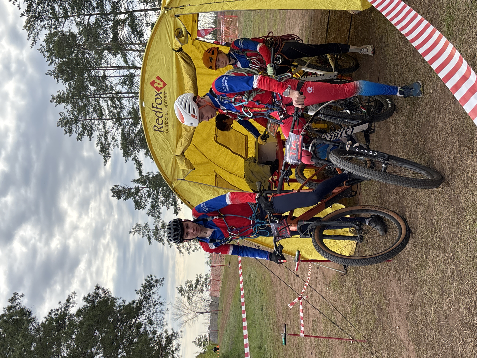 The image shows three people, possibly athletes, with bicycles in a grassy area near a yellow tent. They are wearing helmets and sportswear, with harnesses and climbing equipment. The tent is marked with "RedFox" outdoor equipment branding. A red and white striped tape is visible in the foreground, possibly marking a boundary or course. In the background, there are trees and a cloudy sky. The location is implied to be Orenburg, Russia, based on context not explicitly stated in the image.