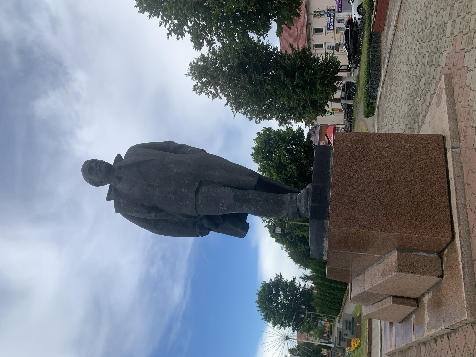 A full-body, low-angle shot captures a large, dark statue of a man standing on a granite pedestal in Pastavy, Belarus. The statue depicts a man wearing a suit and overcoat, with his head turned slightly to his left. The pedestal is a multi-tiered structure, with the main section made of speckled reddish-brown granite. In the background, a paved walkway lined with trees and small hedges leads towards a street with several buildings. Some of the buildings have red tile roofs, and one has signage that includes the Russian word "МОЛОК," meaning "milk." Several cars are parked along the street. The sky above is partly cloudy with patches of blue. In the distance, a large, white, starburst-shaped structure is visible.