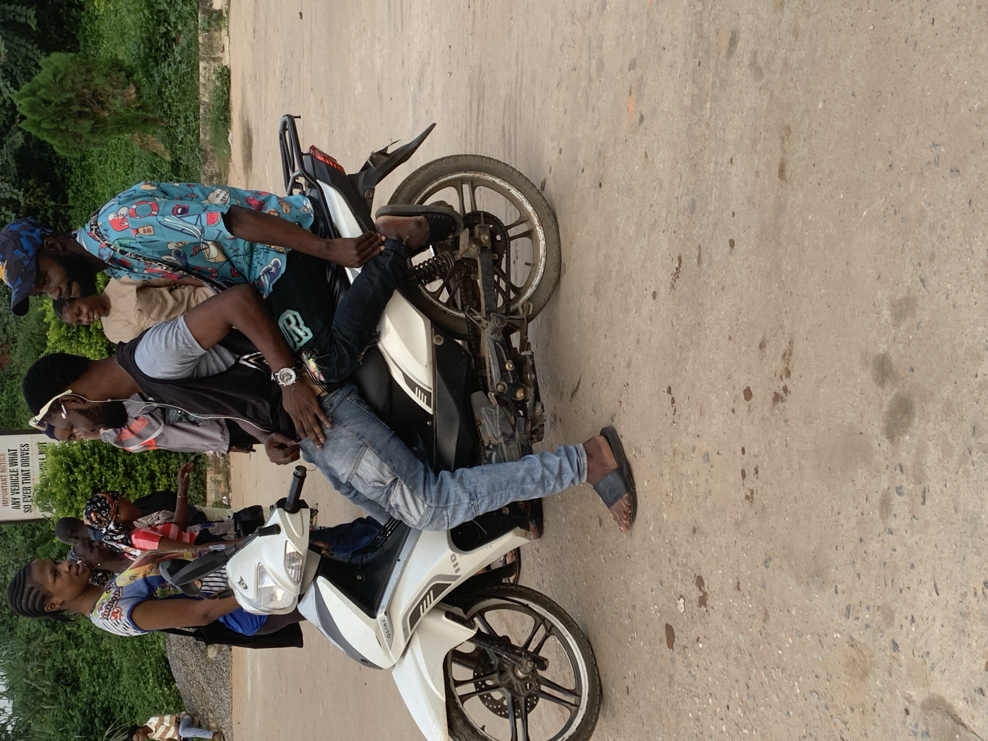 In Shagamu, Nigeria, on a dusty, unpaved street, two men are seated on a white motorcycle. The man in front, wearing a black vest over a grey shirt and blue jeans, has a black watch on his left wrist. He is leaning forward, his hands resting on his thighs. Behind him, a man wearing a blue and black patterned shirt and a blue cap looks to his left. His right arm is extended, resting on the rear of the motorcycle.
A group of people are standing and walking behind the motorcycle. A woman in a colorful t-shirt and blue jeans stands to the left of the motorcycle, looking towards the seated men. Behind her, another woman in a striped t-shirt and headscarf is visible. Further in the background, a man in a light brown shirt sits on a pile of rubble, and a woman in a light brown top stands near some trees. The environment is characterized by dry, cracked earth, scattered small stones, and sparse green foliage in the background, suggesting a warm, dry climate. The lighting indicates it is daytime, possibly overcast due to the diffused light.
