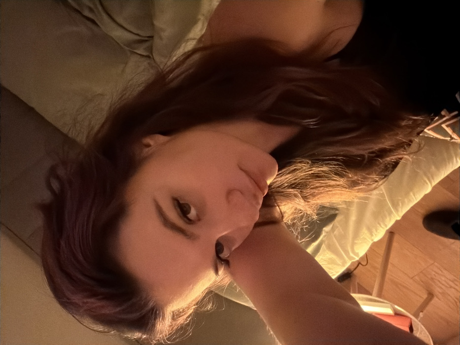 A woman with long brown hair rests her head on her arm. She has fair skin, brown eyes, and a light complexion. The background shows a pillow, bedsheets, and the corner of a wooden table. Soft, warm lighting illuminates the scene. The image is shot from a slightly downward angle.