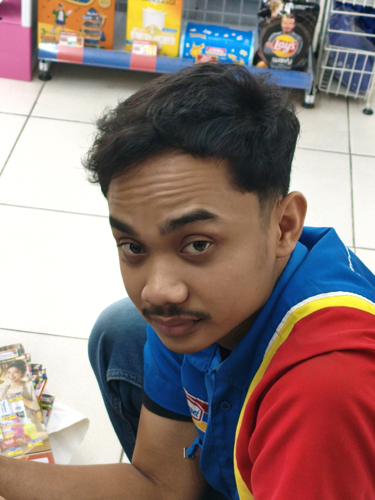 A young man with dark, curly hair and a mustache is crouching on a tiled floor in what appears to be a convenience store. He is wearing a blue shirt with red and yellow trim and blue jeans. He is looking directly at the camera with a slight smile. In the background, shelves are stocked with various products, including snacks and beverages. The lighting is bright and even, suggesting an indoor commercial setting. The floor is tiled in a grid pattern. There are some products on a shelf to the left of the man, including items with images of women and food. A rack with a bag of Lays chips and other bagged snacks is visible behind him.