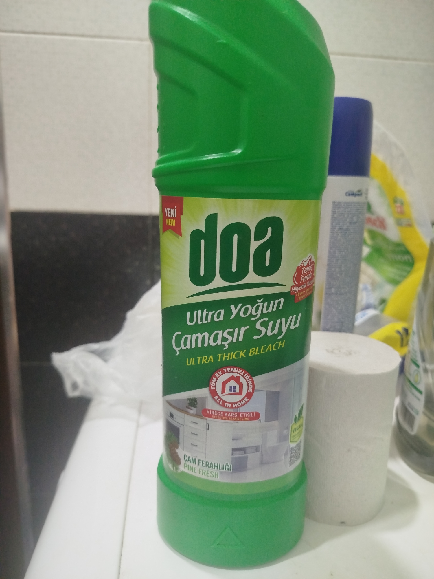 The image shows a close-up of a green bottle of "doa Ultra Yoğun Çamaşır Suyu" (Ultra Thick Bleach) standing on a white surface. To the right of the bottle, a roll of white toilet paper is visible, and behind it, another cleaning product bottle with a blue cap can be seen. The background is a blurred mix of white tiles and dark surfaces. The scene appears to be indoors, possibly in a bathroom or kitchen, given the presence of cleaning supplies. There are no people or animals in the image, and no specific geographical markers are evident that would indicate its location in Batumi, Georgia.