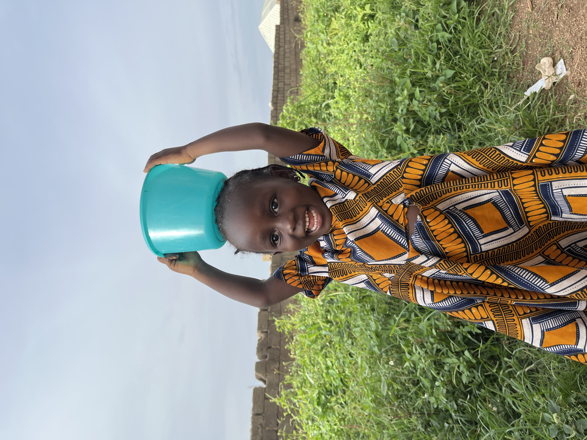 In Rafin Par, Nigeria, a young girl with dark skin and braided hair smiles widely, holding a turquoise bowl over her head. She is wearing a vibrant, patterned dress in orange, white, and blue. The dress features geometric designs and a repeating wave-like motif. Her arms are raised, supporting the bowl.
The background shows a lively scene: lush green grass and foliage are abundant, suggesting a natural or outdoor setting. A low brick wall is visible in the middle ground, and beyond that, the edge of a white-tiled roof of a building can be seen, hinting at a settlement. The sky above is a soft, pale blue, indicating it's daytime, likely under clear or partly cloudy weather conditions. The ground appears to be dirt and grass. The overall impression is one of joy and playfulness, captured in a natural, possibly rural, environment. There is no visible text in the image.