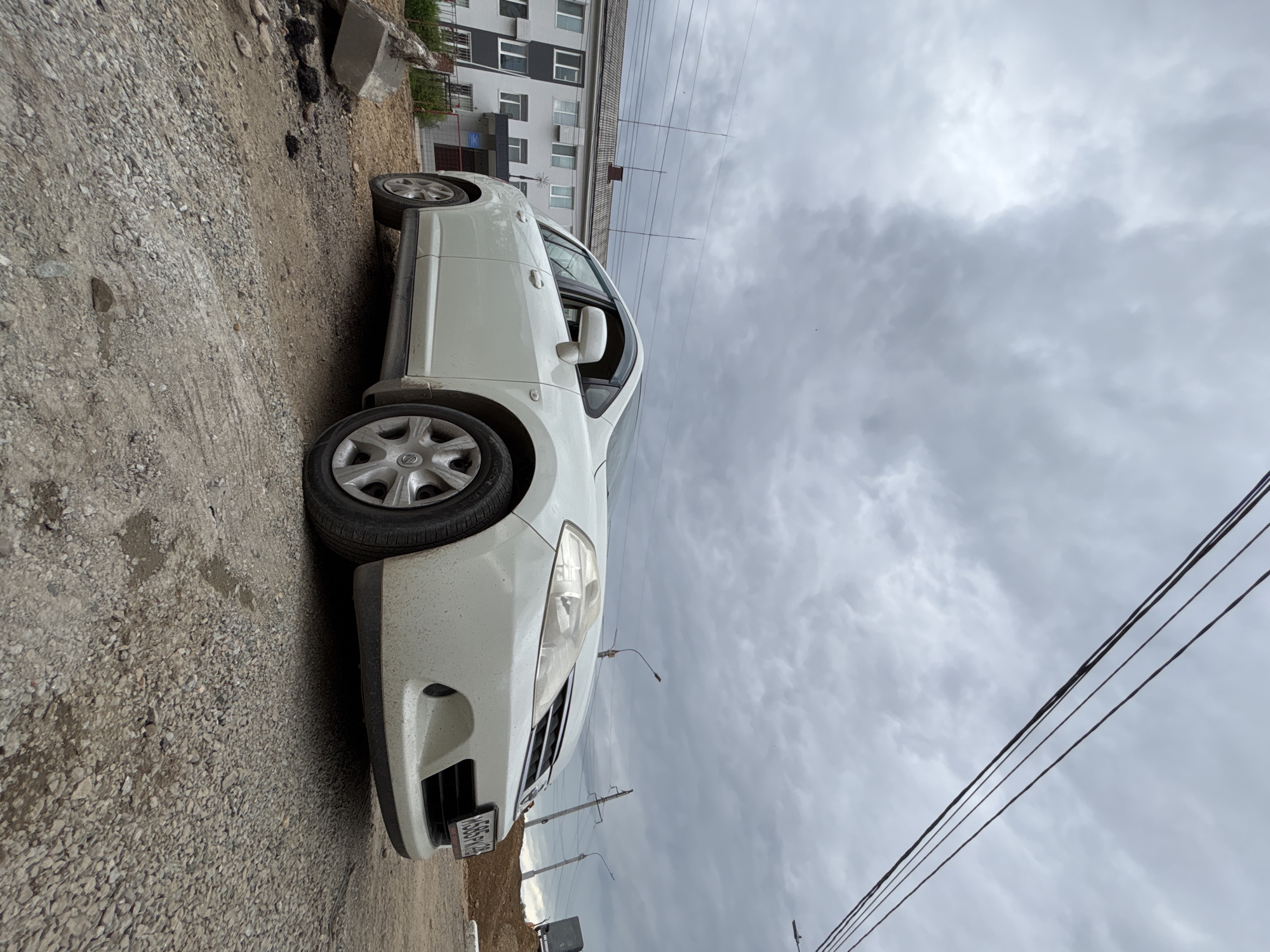 A white hatchback is parked on a gravel surface in front of a light-colored, multi-story building with dark accents. The building has numerous windows. Overhead, power lines crisscross the cloudy sky. The car's license plate is partially visible, displaying Cyrillic characters and numbers. Debris, including a concrete fragment, is scattered on the ground near the car. The scene appears to be an outdoor area in an urban or suburban setting. The presence of Cyrillic on the license plate suggests a Russian location. Given the specific license plate format, it is highly likely to be Blagoveshchensk, Russia.