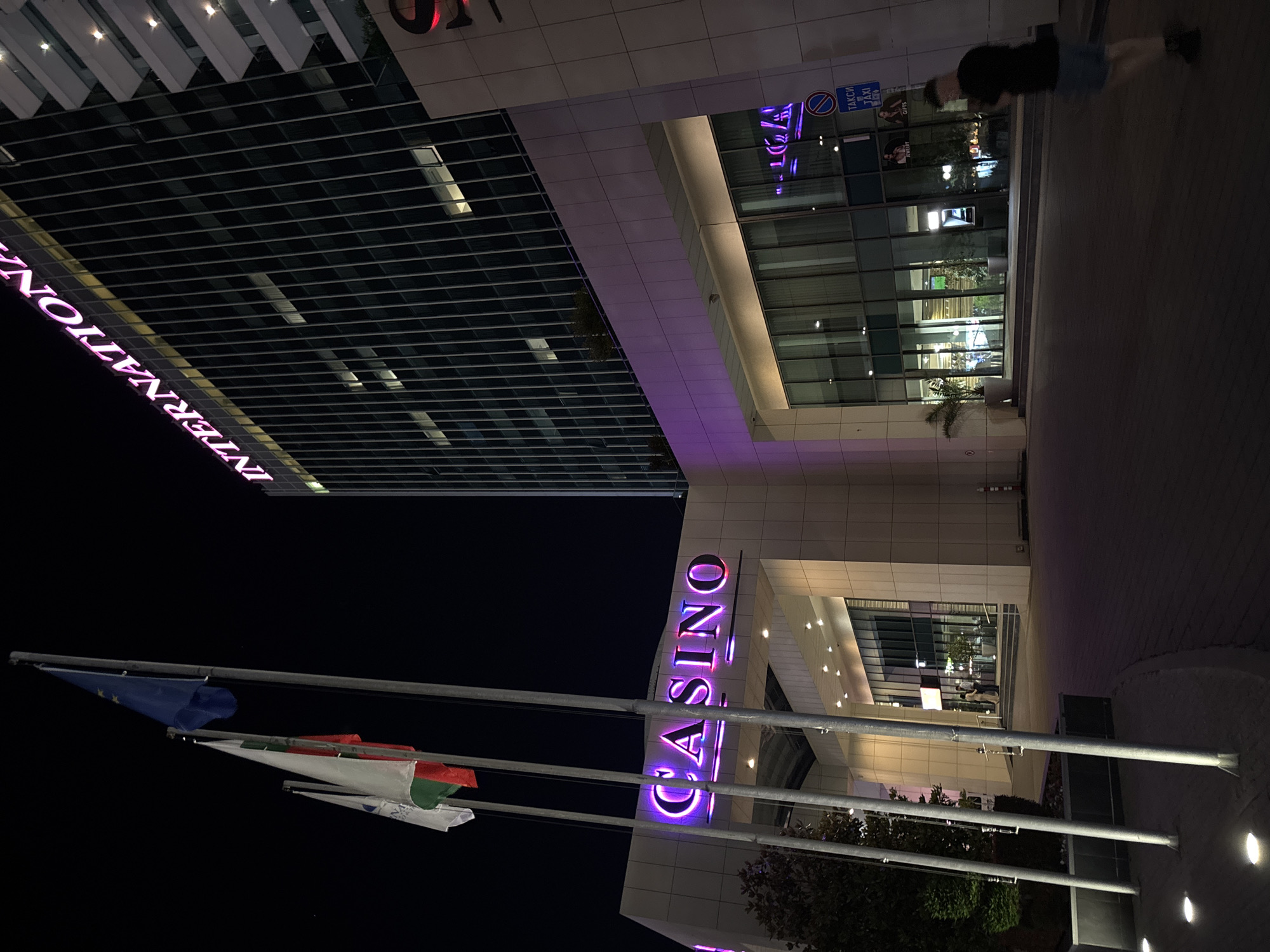 The image captures the "International" hotel and casino located in Varna, Bulgaria, at night. The imposing modern architecture of the hotel building is illuminated, with the word "INTERNATIONAL" prominently displayed in neon pink lettering across its upper facade. Below, the entrance to the casino is marked by a bright pink neon sign spelling out "CASINO". Several flags, including the European Union flag and Bulgarian flags, are planted on flagpoles in front of the casino entrance. The scene is well-lit, with artificial lights highlighting the building's features and the surrounding walkway. A few people are visible, including one individual jogging on the sidewalk to the right of the frame and others blurred in the background near the casino entrance. The ground is paved, and trees and planters are strategically placed around the entrance. A blue taxi sign is visible on the building's facade, indicating the availability of taxi services. The overall atmosphere is one of a vibrant urban night scene, typical of a casino and hotel complex.