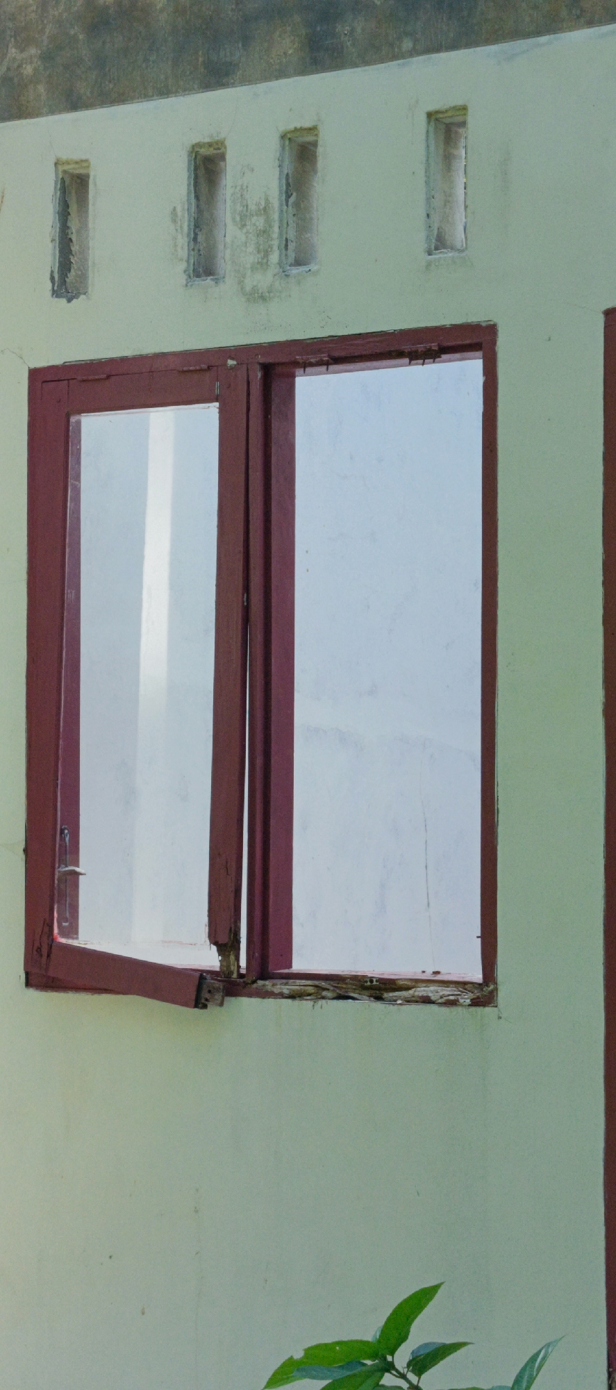 The image shows a weathered, pale green wall of a building in Pashiebeutong, Indonesia. There is a large, double-paned window with dark red wooden frames. The left pane of the window is open, revealing a glimpse of the interior or sky beyond. The wood of the window frame appears worn and possibly damaged in places. Above the window, there are three small, rectangular openings in the wall, also with weathered frames. The wall itself shows signs of age, with some discoloration and what appear to be faint cracks. In the lower right corner of the image, the tips of green leaves from a plant are visible, suggesting some vegetation near the building.