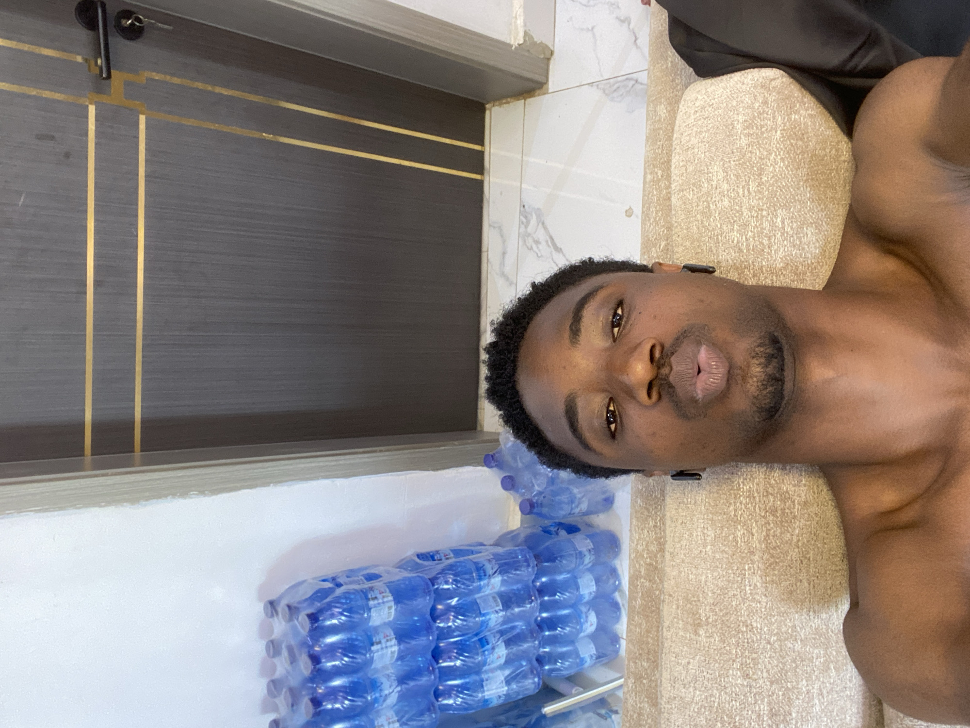 A shirtless dark-skinned man with short, curly black hair and a goatee is lying on a beige upholstered surface. He is looking at the camera with his mouth slightly open. To the left of the man is a stack of bottled water in blue plastic packaging. Above the bottled water is a door with gray paneling and gold accents. The image appears to have been taken indoors. There is no indication of Ibadan, Nigeria.