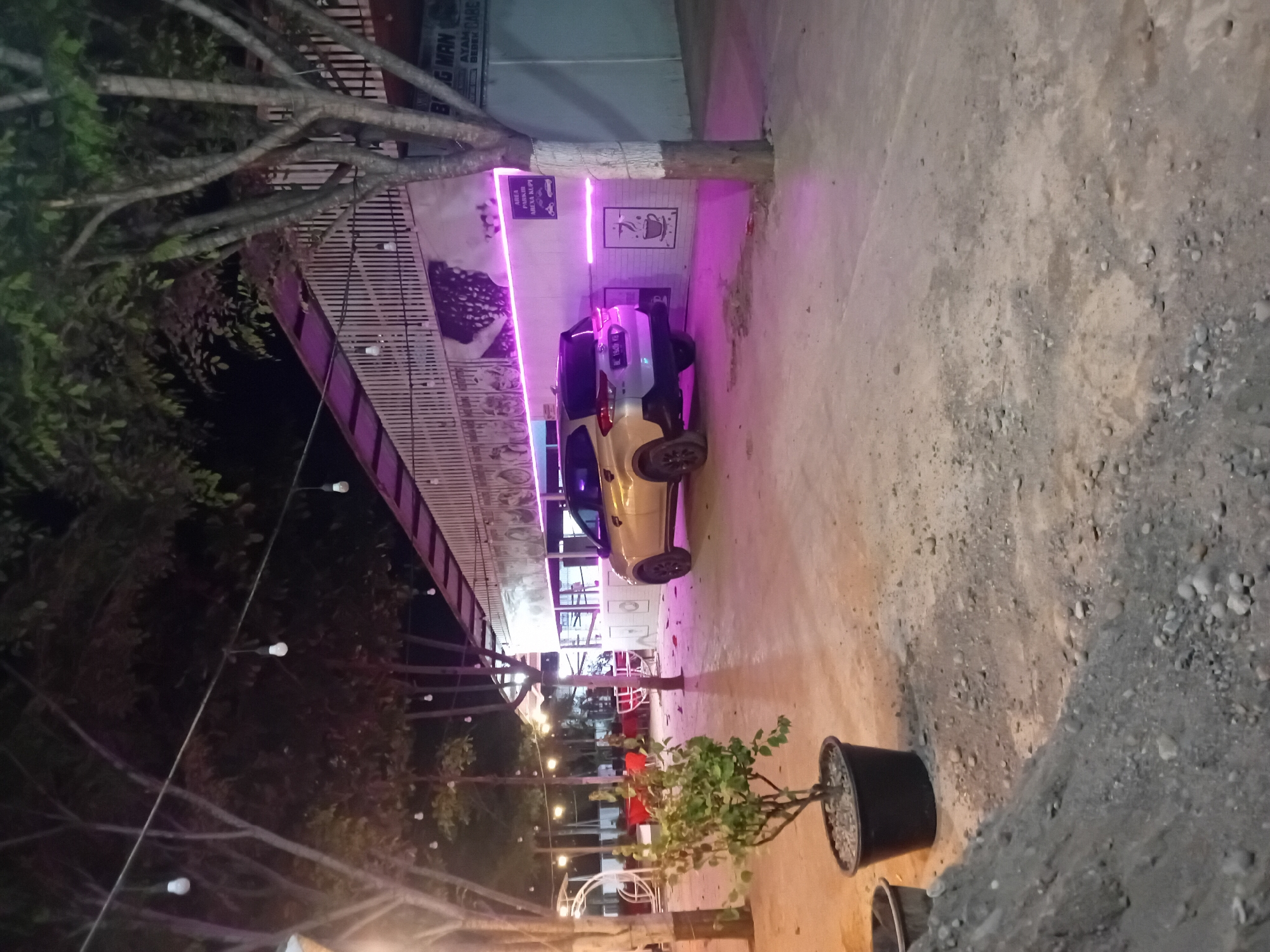 The image depicts an outdoor scene at night, likely a roadside establishment in Cotkumbang, Indonesia, characterized by a dirt and gravel ground surface. A light-colored car is parked on the right side of the frame, facing towards the left. Behind the car is a building with a white slatted facade, illuminated by pink neon lights along its base. A sign with text, partially obscured by a tree, is visible on the building. In the foreground, a potted plant with green foliage sits on the ground to the left of the parked car. The background features trees with visible branches and leaves, strung with decorative lights. The overall ambiance suggests a casual dining or gathering spot.