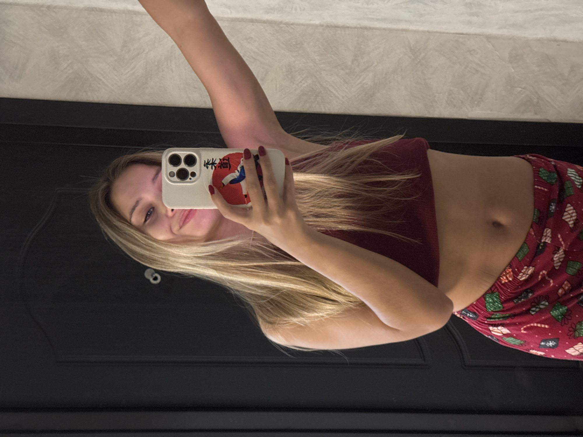 A young woman with blonde hair takes a selfie in a mirror. She is wearing a burgundy crop top and red patterned pajama bottoms. Her right arm is extended, holding a white smartphone with a case depicting a martial artist. The background shows a dark door on the left and a textured wall on the right. The scene appears to be indoors, possibly in a residential setting in Chaikovsky, Russia, though this cannot be definitively confirmed by the visual information alone.
